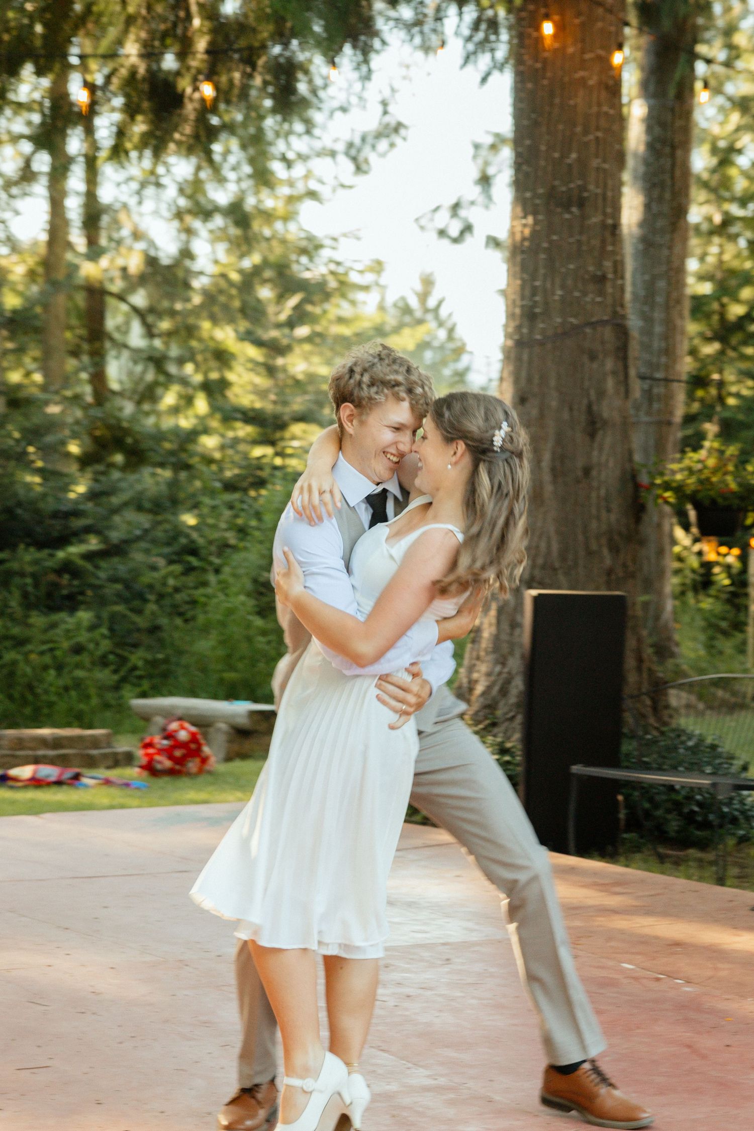 A joyful newlywed couple shares a romantic first dance at their outdoor wedding reception, surrounded by trees and warm string lights.