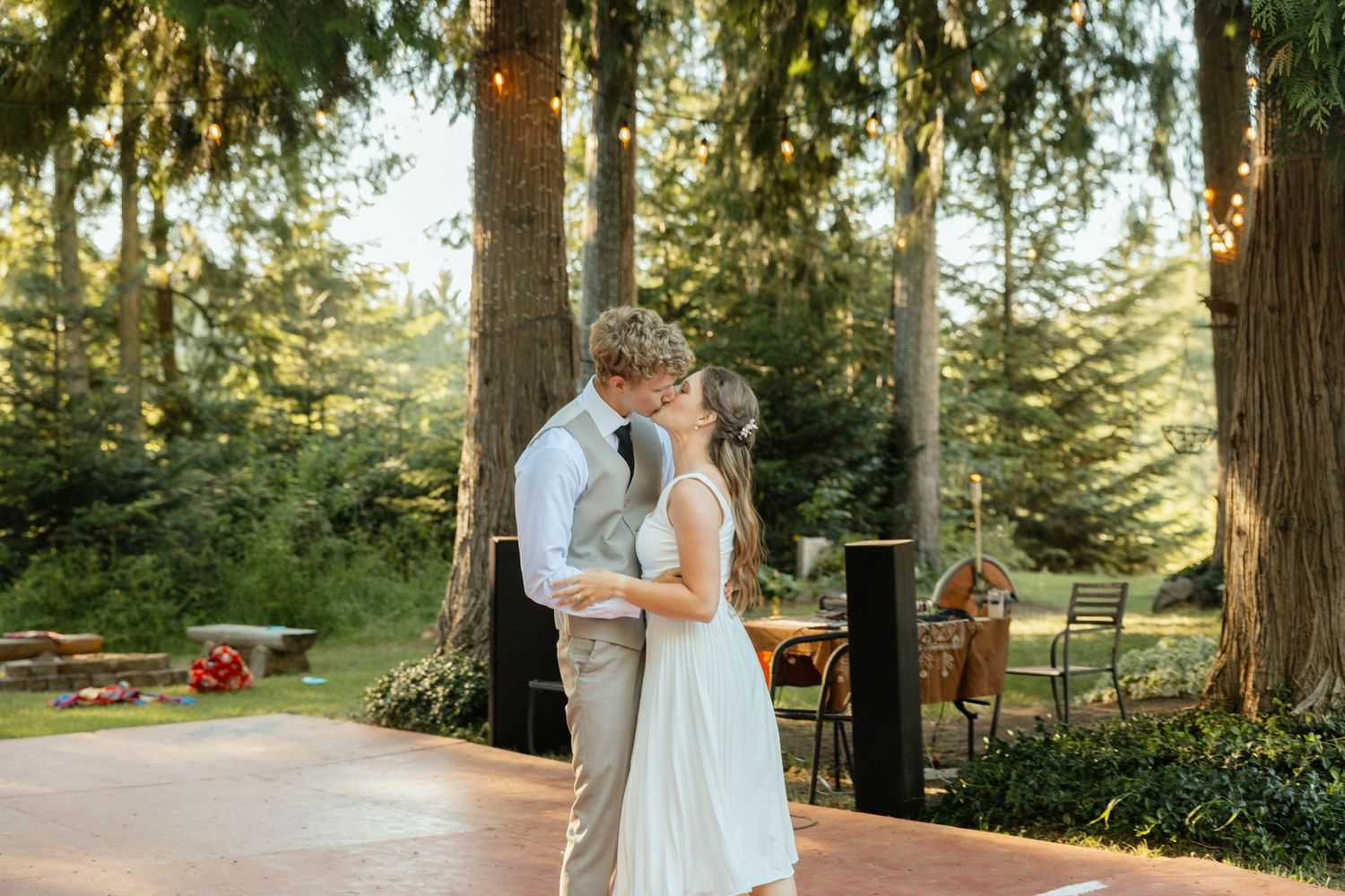 A romantic kiss after the bride and groom share their first dance beneath trees, with soft string lights and warm evening light.