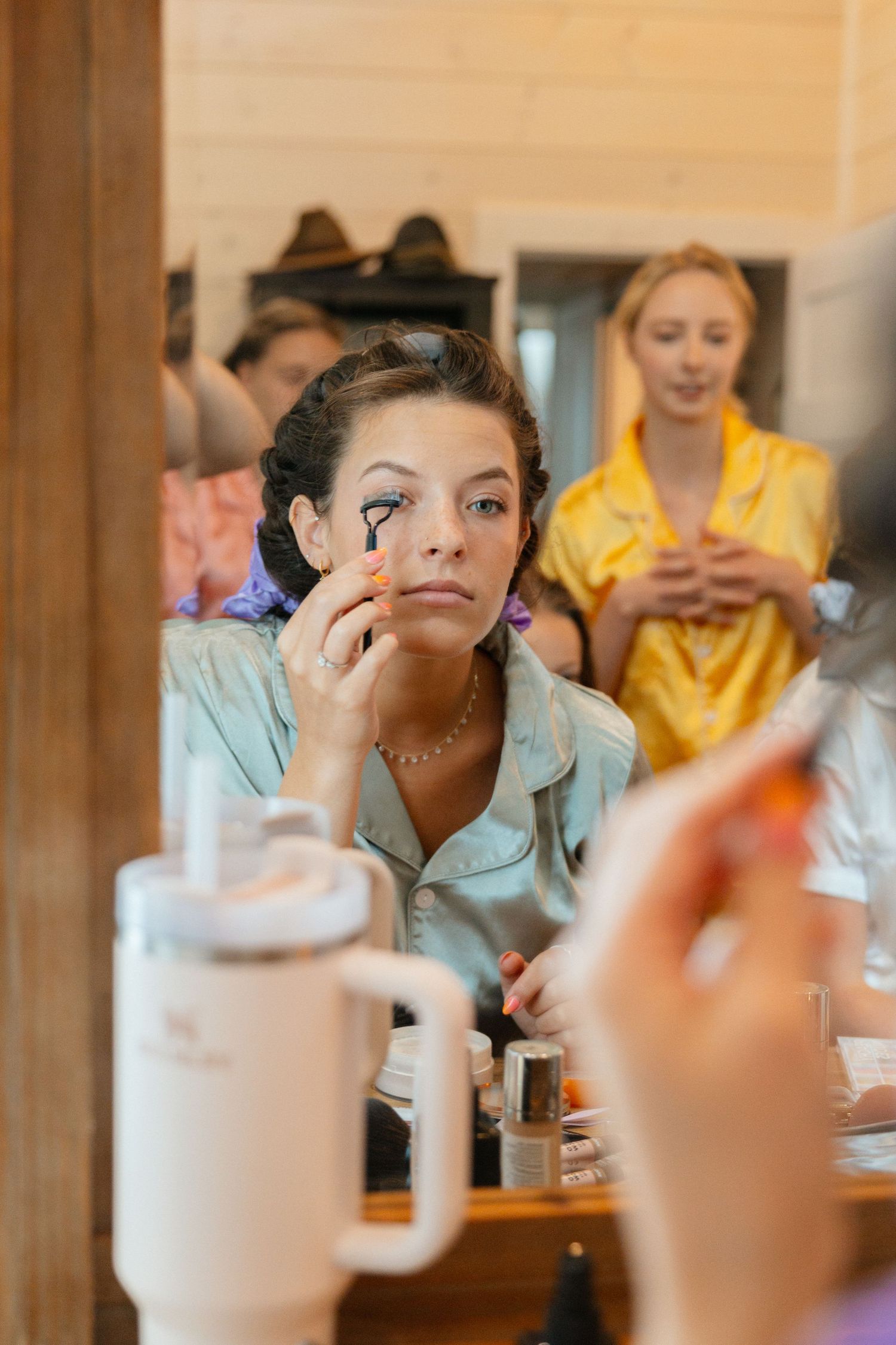 Bridesmaid in silk blue set curling eyelashes in front of a mirror with colorful bridesmaids in the background getting ready for the wedding