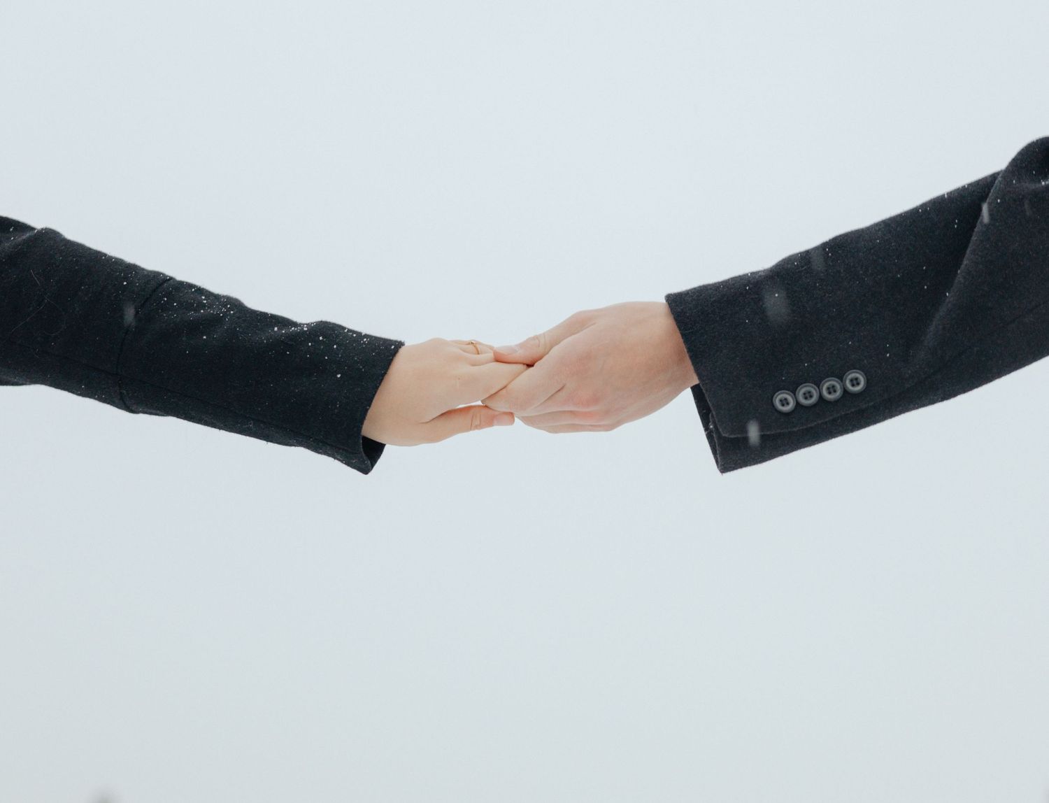 Close-up of a couple holding hands in the snow, symbolizing love, connection, and commitment against a minimalist winter sky backdrop