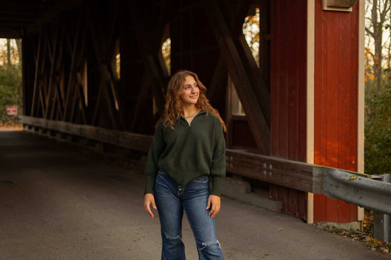 Confident young woman standing near a rustic red covered bridge, bathed in golden light, evoking warmth, strength, and timeless charm.