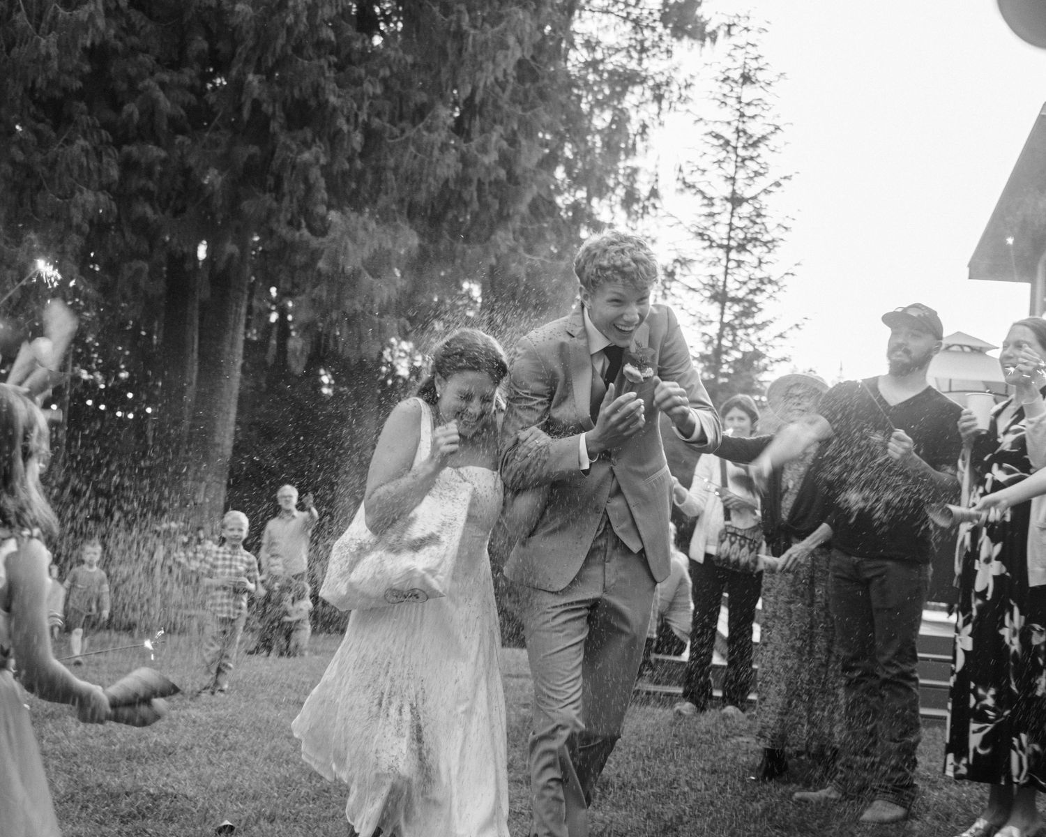 A black-and-white scene of a bride and groom laughing and running as they exit their forest wedding reception, surrounded by joyful guests.