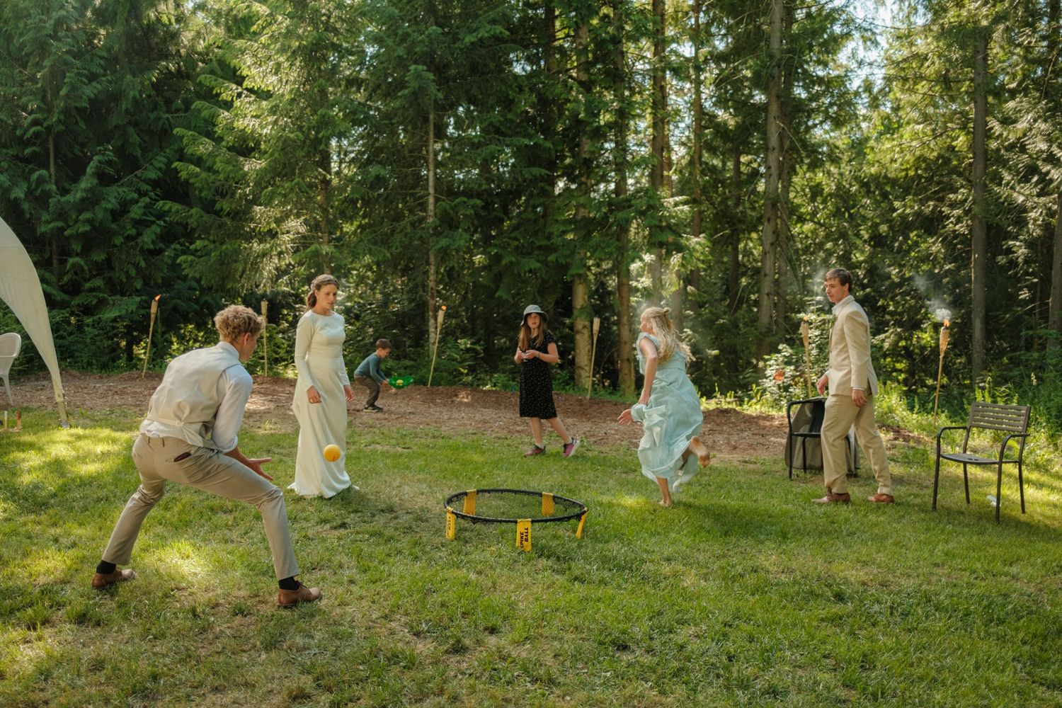 A wedding party enjoys a lively spikeball game in a sunlit forest clearing, with the bride in white and guests sharing a festive moment.