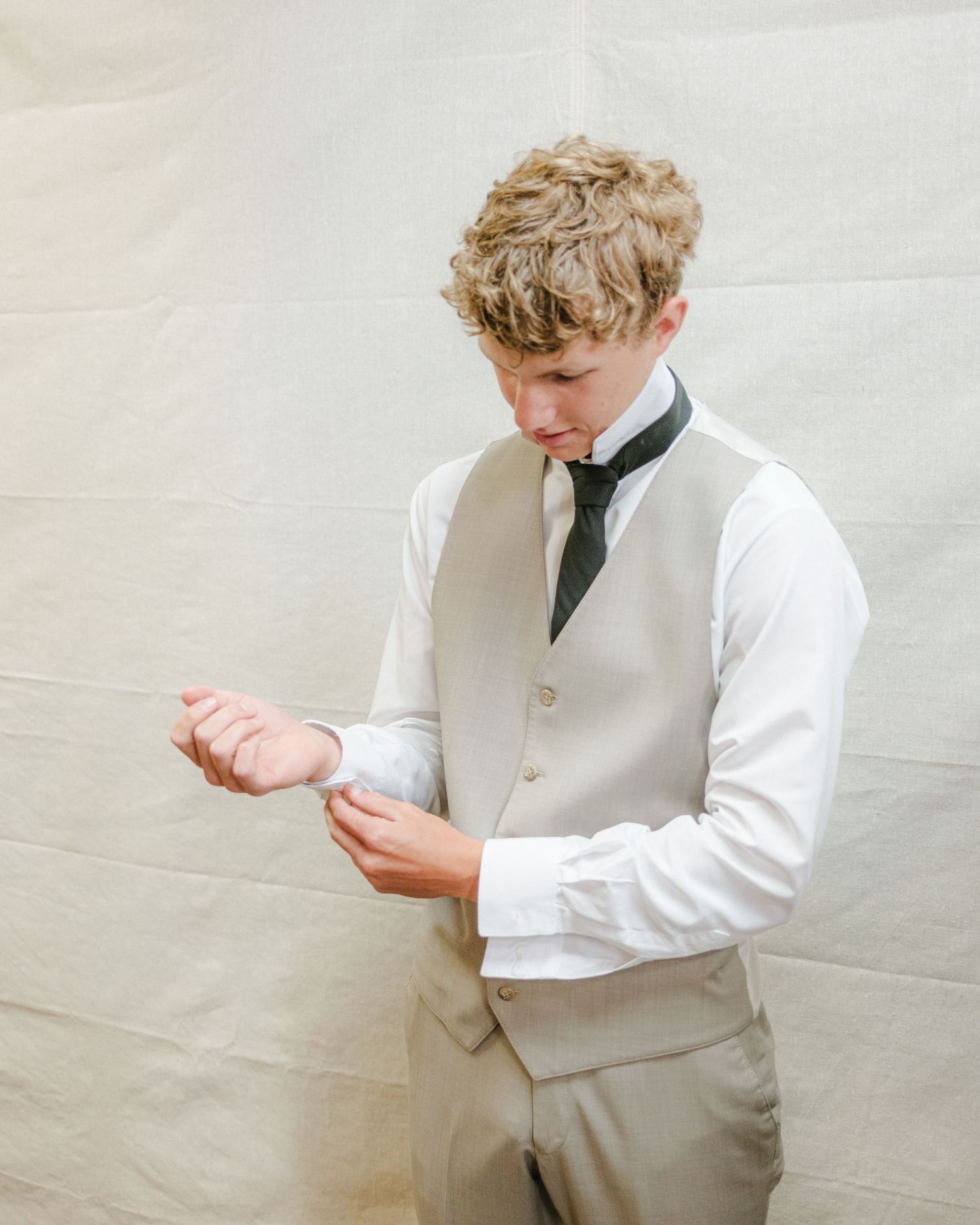 A groom getting ready in a light gray vest and tie adjusts his cuffs, exuding timeless elegance against a soft, draped backdrop.