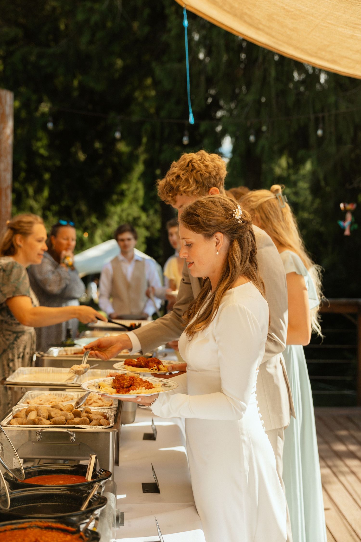 A bride in a white dress dishes up food at a wedding buffet under a tent, surrounded by guests and a lush forest backdrop.