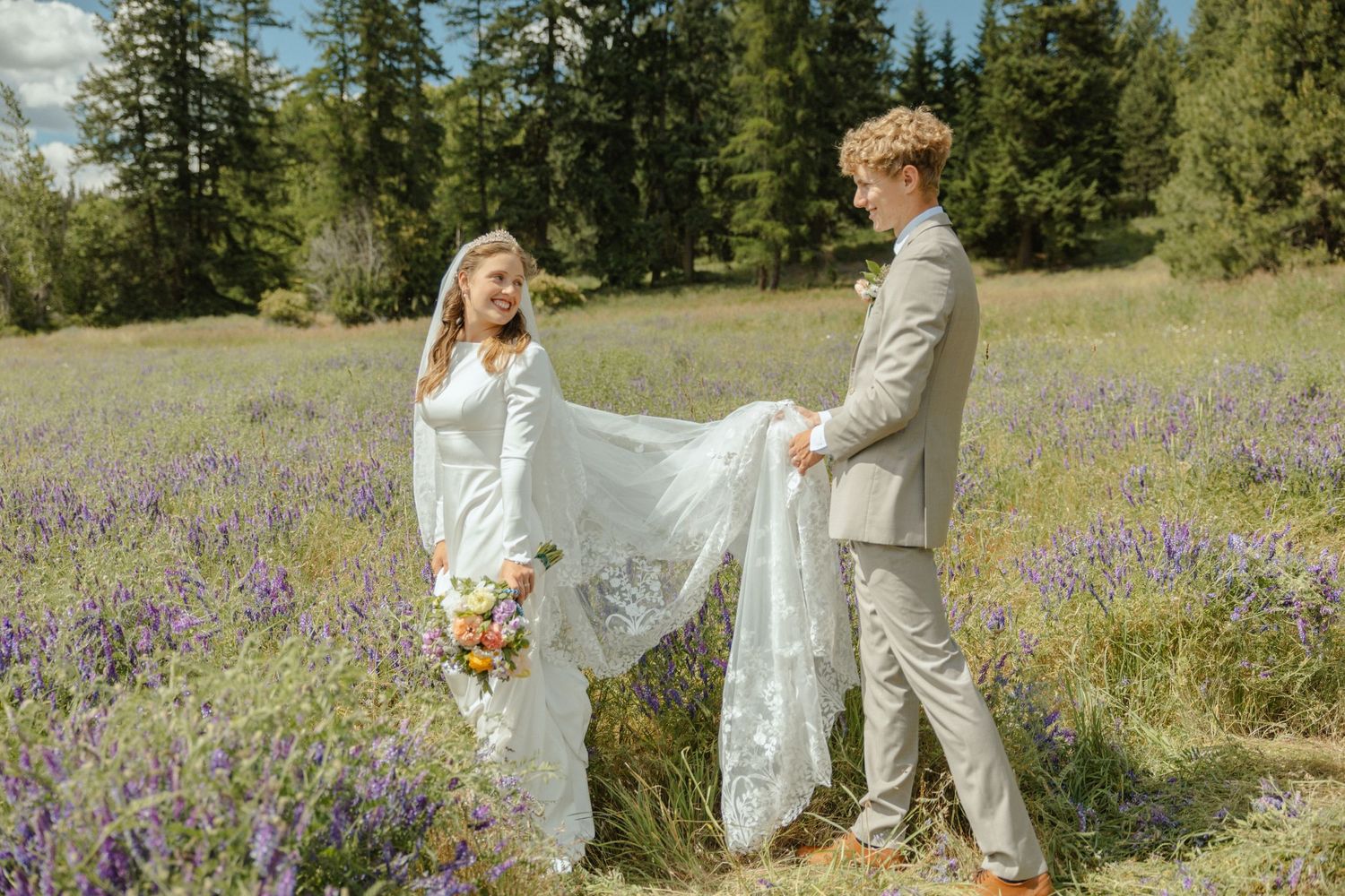 A bride in a flowing gown with a veil and a groom in a light suit stand in a wildflower field, holding her train with trees behind them.