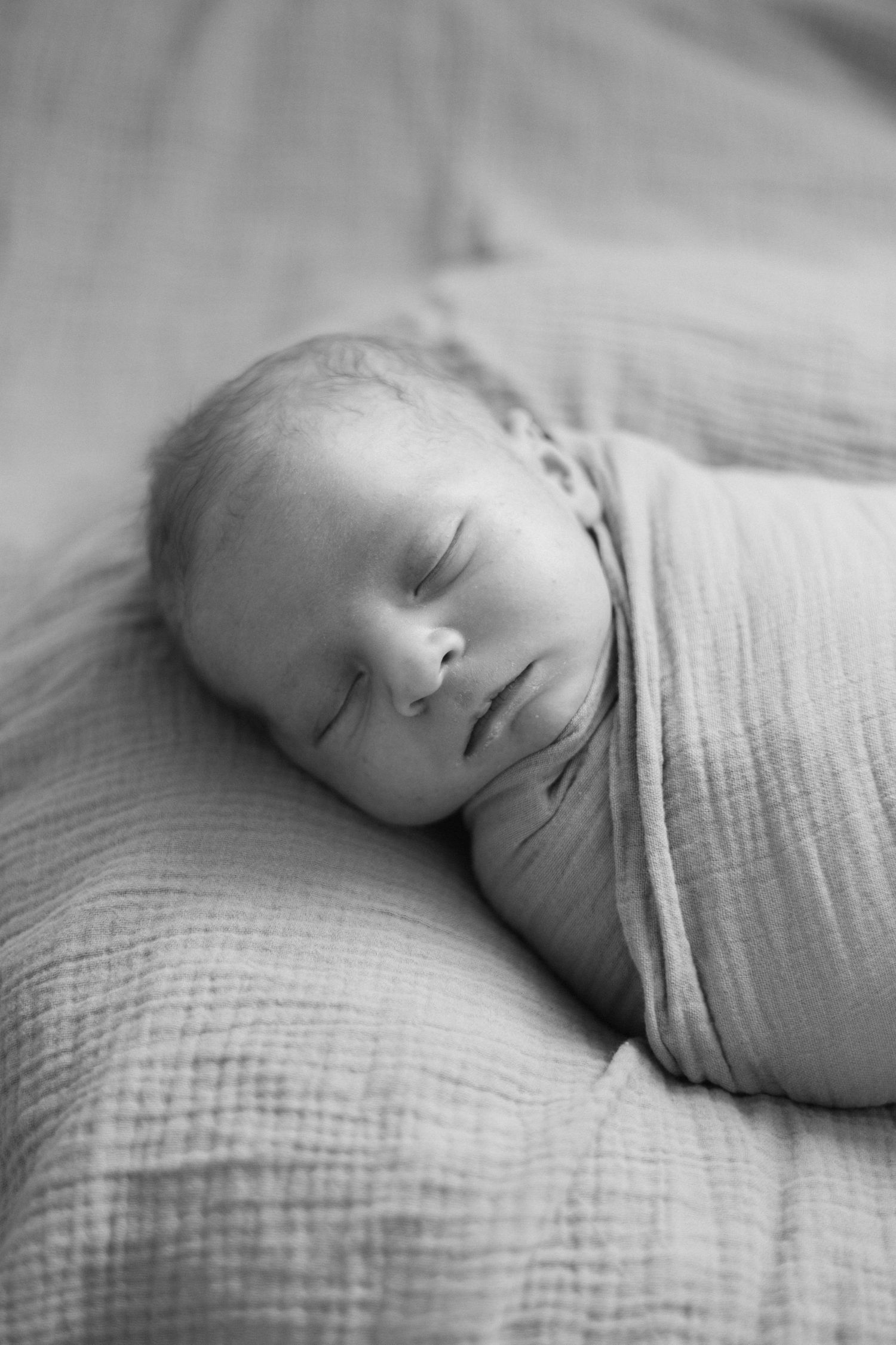 Black and white photo of a newborn baby peacefully swaddled in a soft blanket, resting comfortably on a textured pillow.