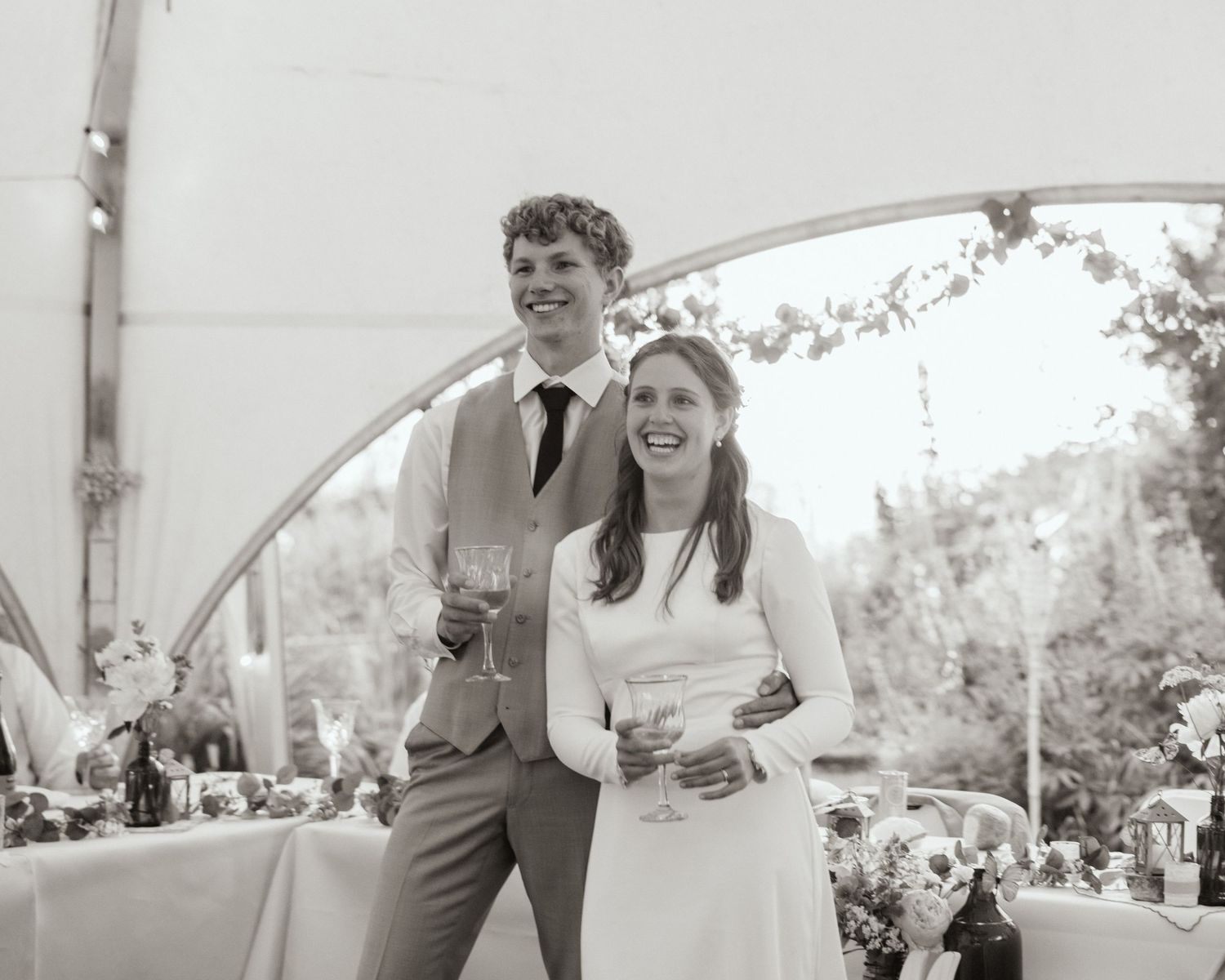 A bride in a long-sleeved gown and groom in a suit share a toast under a tent, smiling warmly with decorated table and greenery behind them