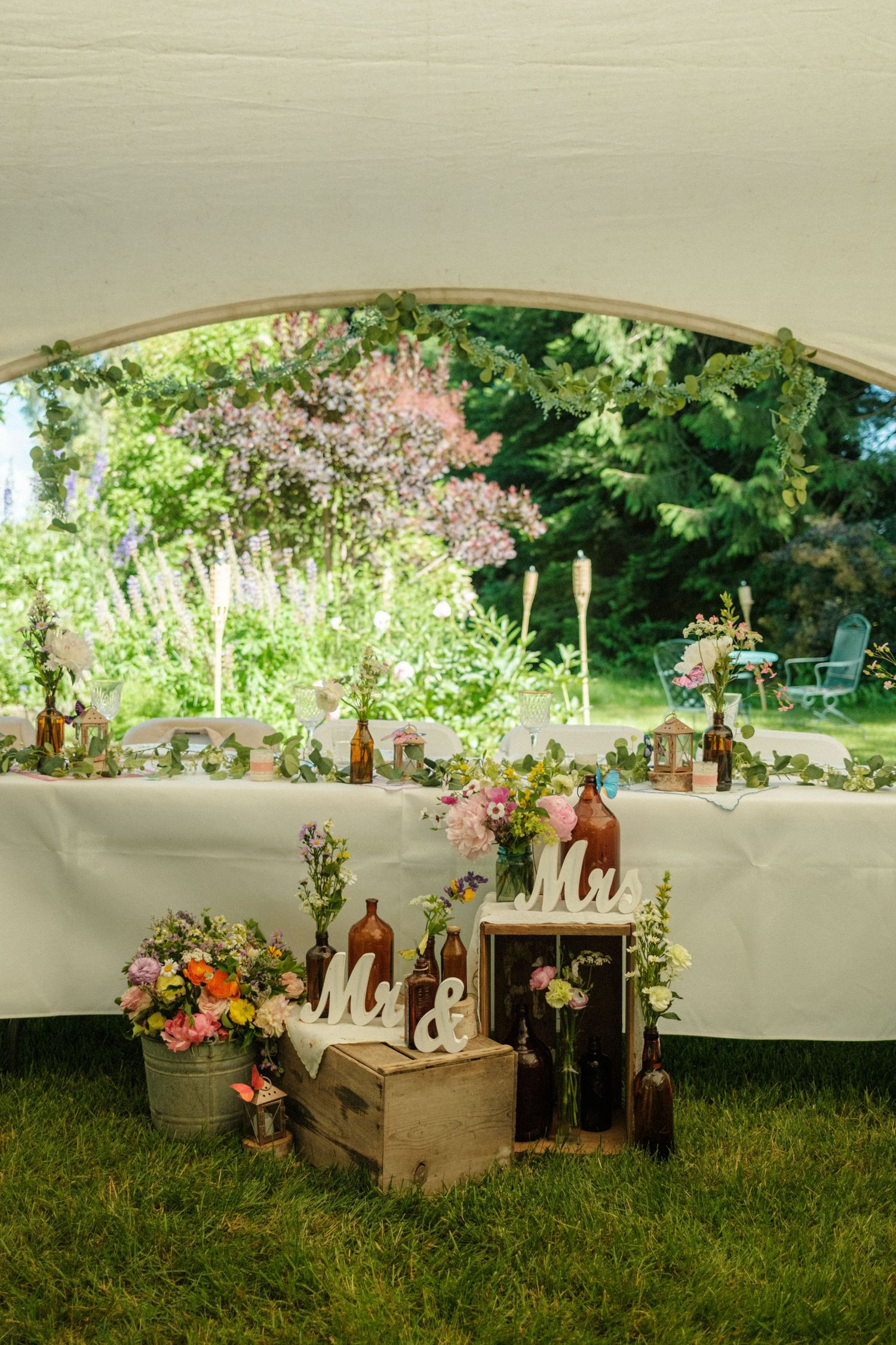 A beautifully decorated wedding table under a tent, adorned with flowers, bottles, and 