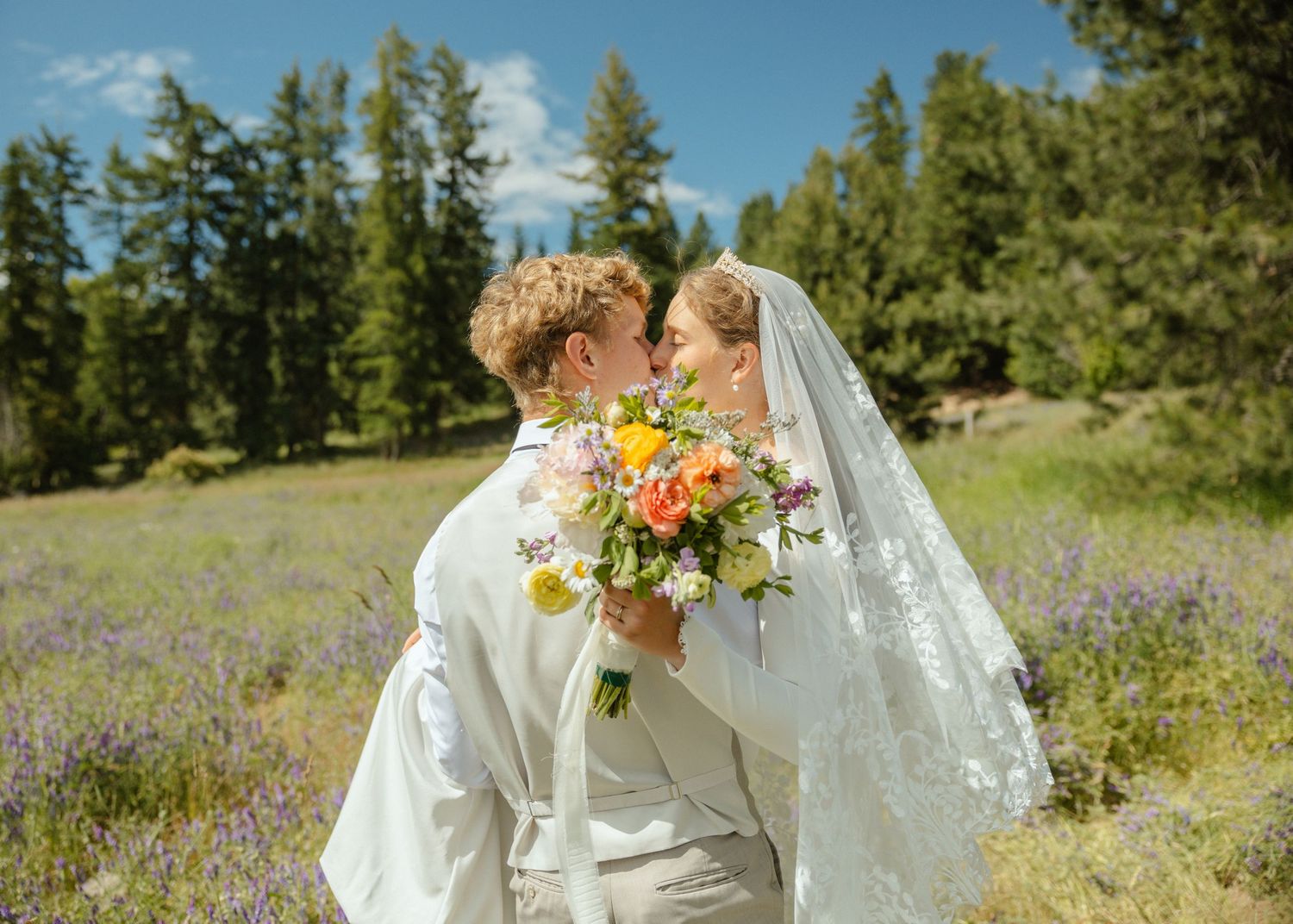 A Groom holding his bride in a sunlit meadow of purple flowers with trees in the distance.The bride is holding a beautiful colorful bouquet