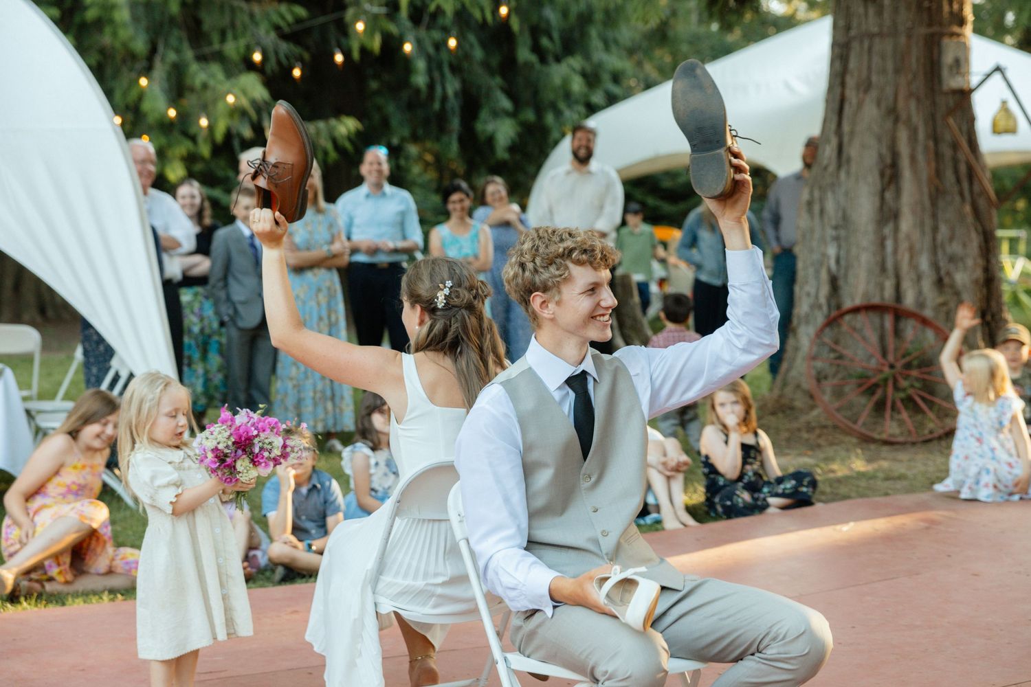 Bride and groom play the shoe game at their outdoor reception, surrounded by guests, children, and trees with string lights glowing above.