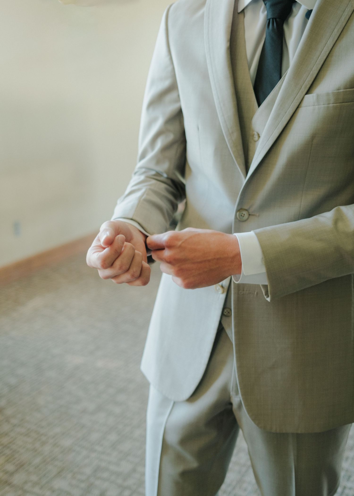 A groom in a light gray suit adjusts his suit cuffs with care, showcasing elegance in a softly lit indoor space.