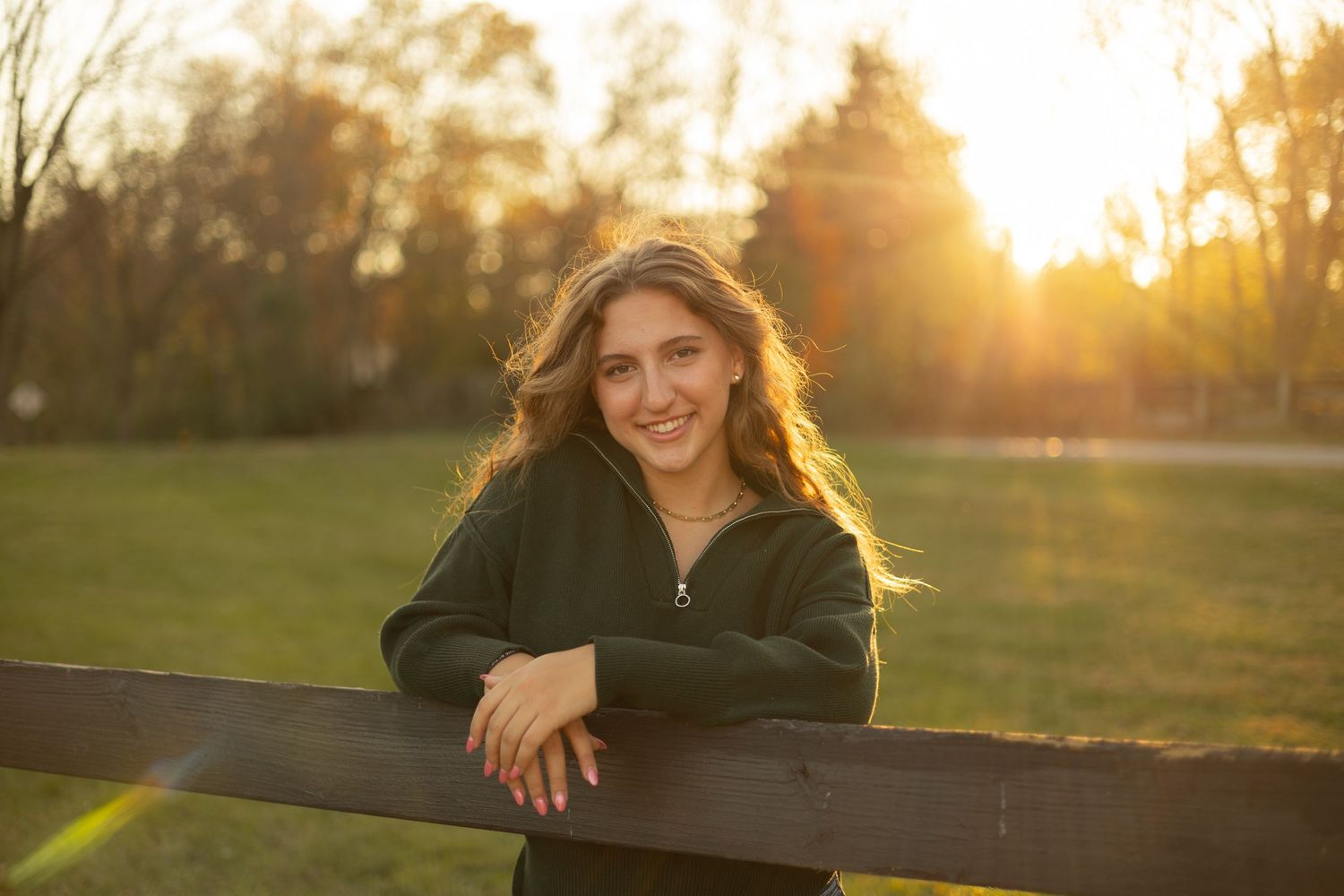 A woman in a dark green sweater leans on a wooden fence, smiling warmly, with a golden sunset and lush greenery in the background.