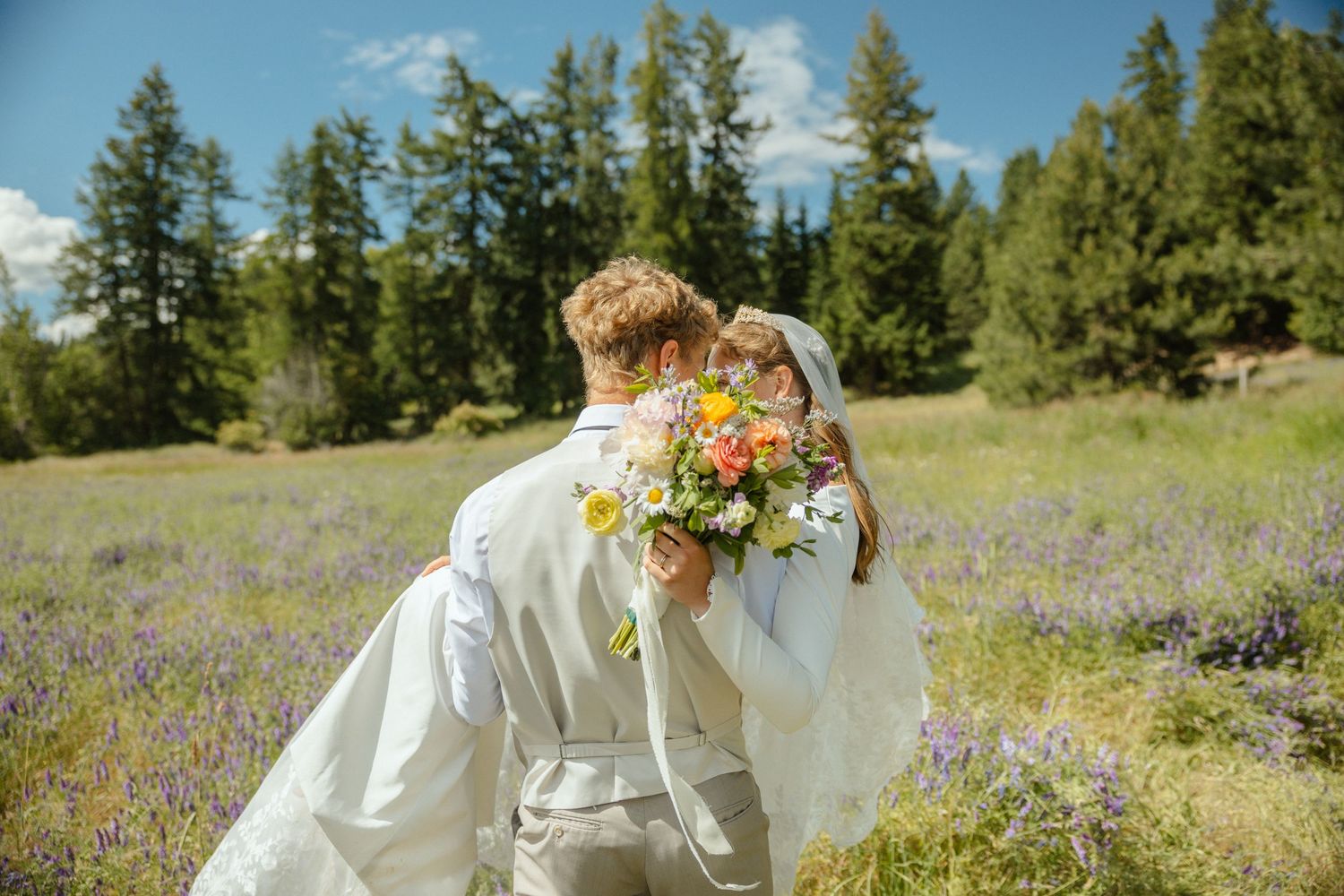A Groom holding his bride in a sunlit meadow of purple flowers with trees in the distance.The bride is holding a beautiful colorful bouquet