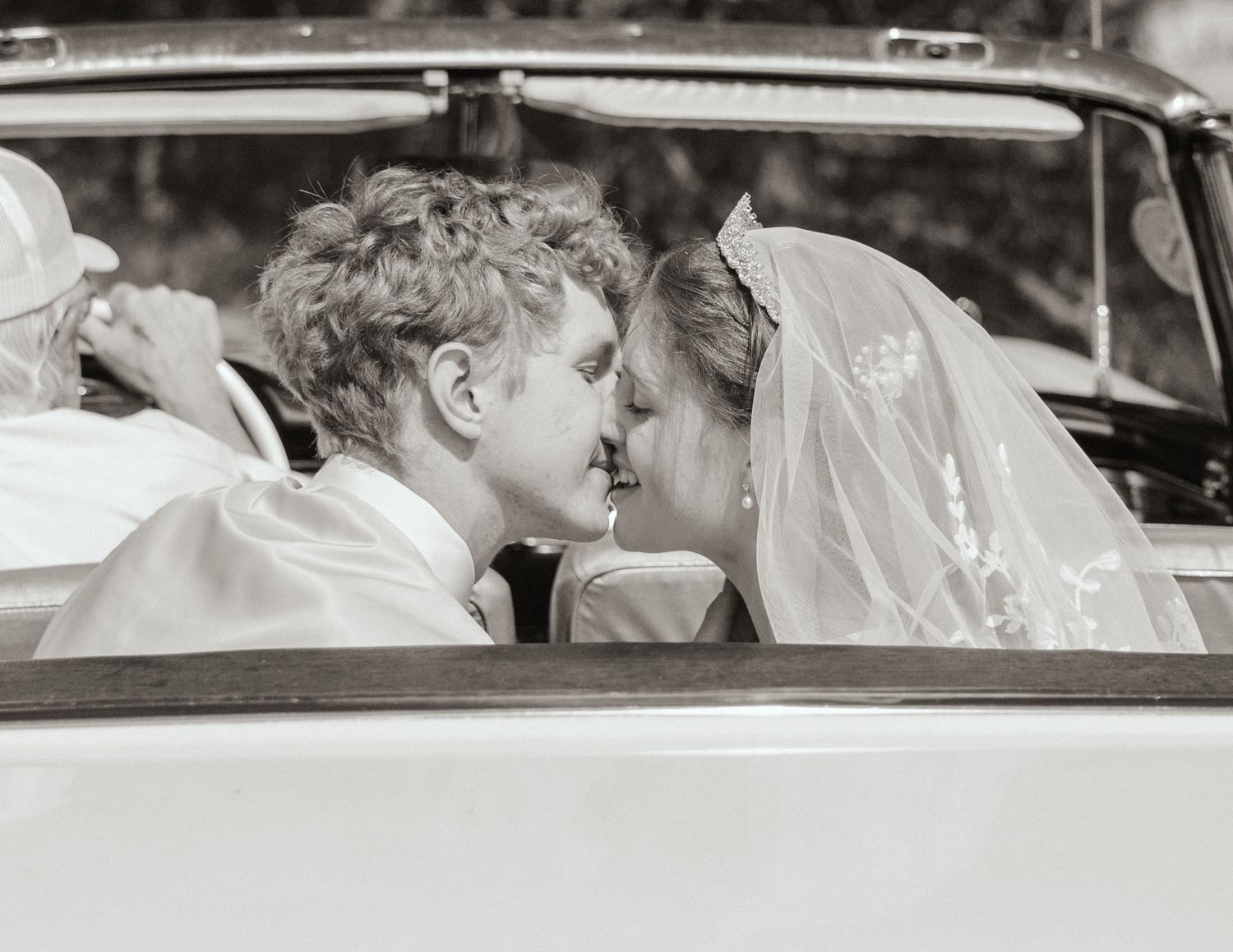 Black-and-white image of a just-married couple in the back of a classic convertible, about to kiss and smiling with so much joy.