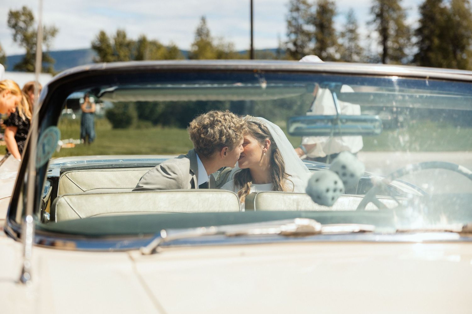A bride and groom share a kiss in a vintage convertible, her veil flowing, set against a scenic backdrop of trees and open sky. Timeless.