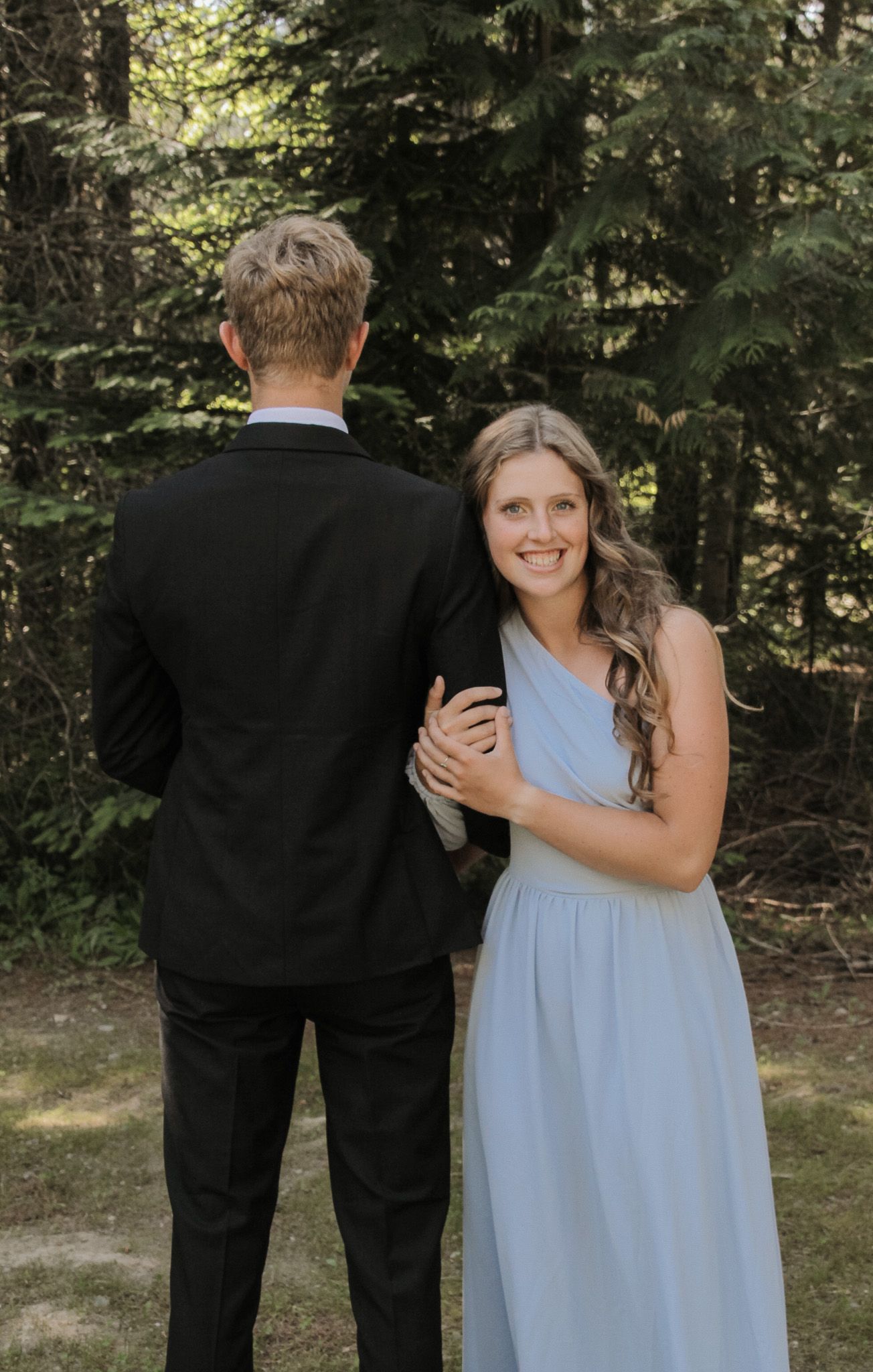 A couple shares a tender moment in a forest: he in a sleek black suit, she in a flowing light blue gown, her smile glowing with joy.