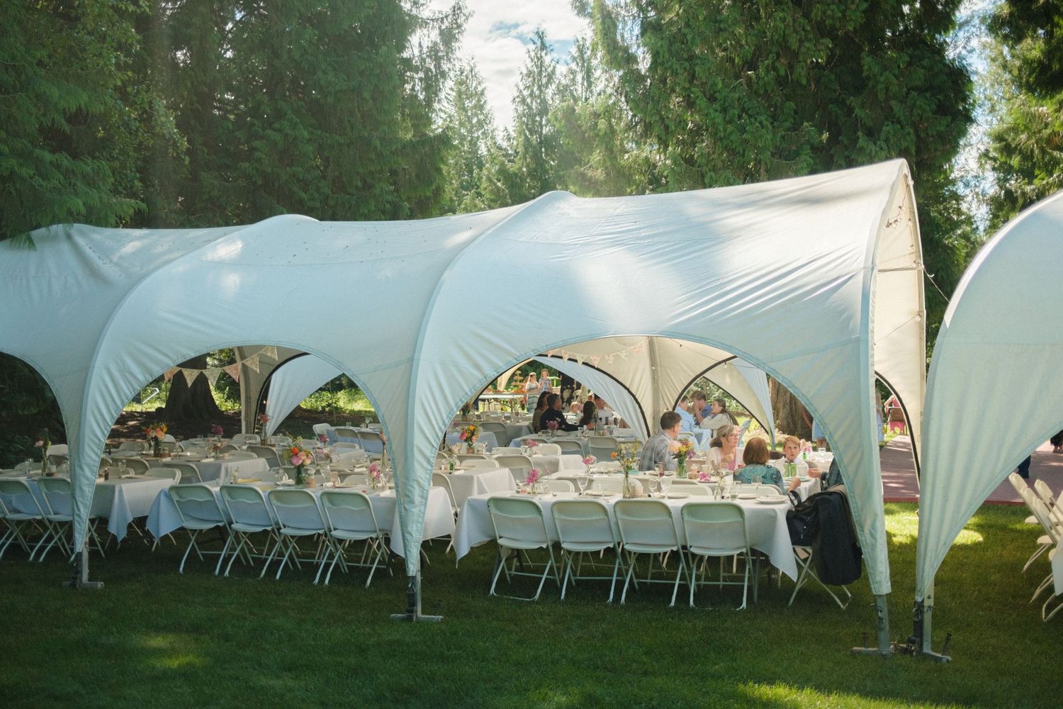 Elegant outdoor wedding reception setup under white canopy tents, featuring beautifully decorated tables surrounded by guests and big trees