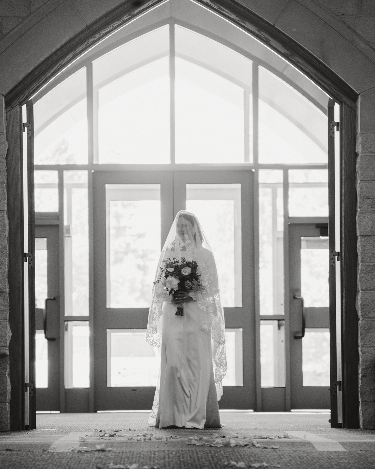 Bride in a flowing gown and veil stands in a church doorway, holding a bouquet, framed by arched windows in a timeless black-and-white image