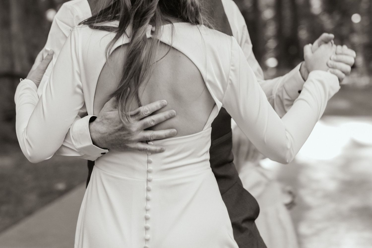A black-and-white image of a father and daughter dancing, her backless white gown with elegant buttons, his hands gently holding hers.
