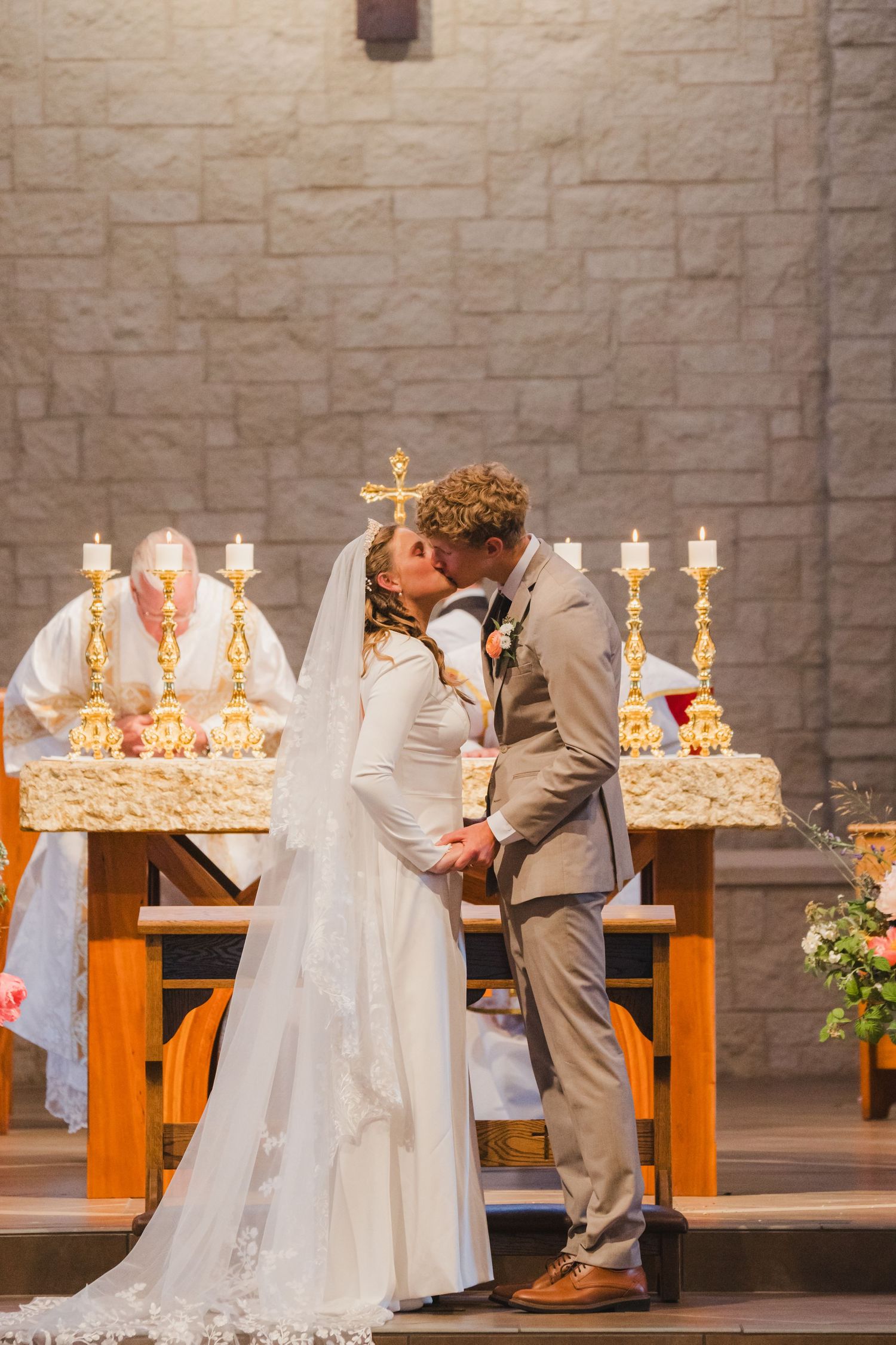 Bride and groom share first kiss at the altar in a stone-walled church, with a priest and lit candelabras creating a sacred, elegant scene.