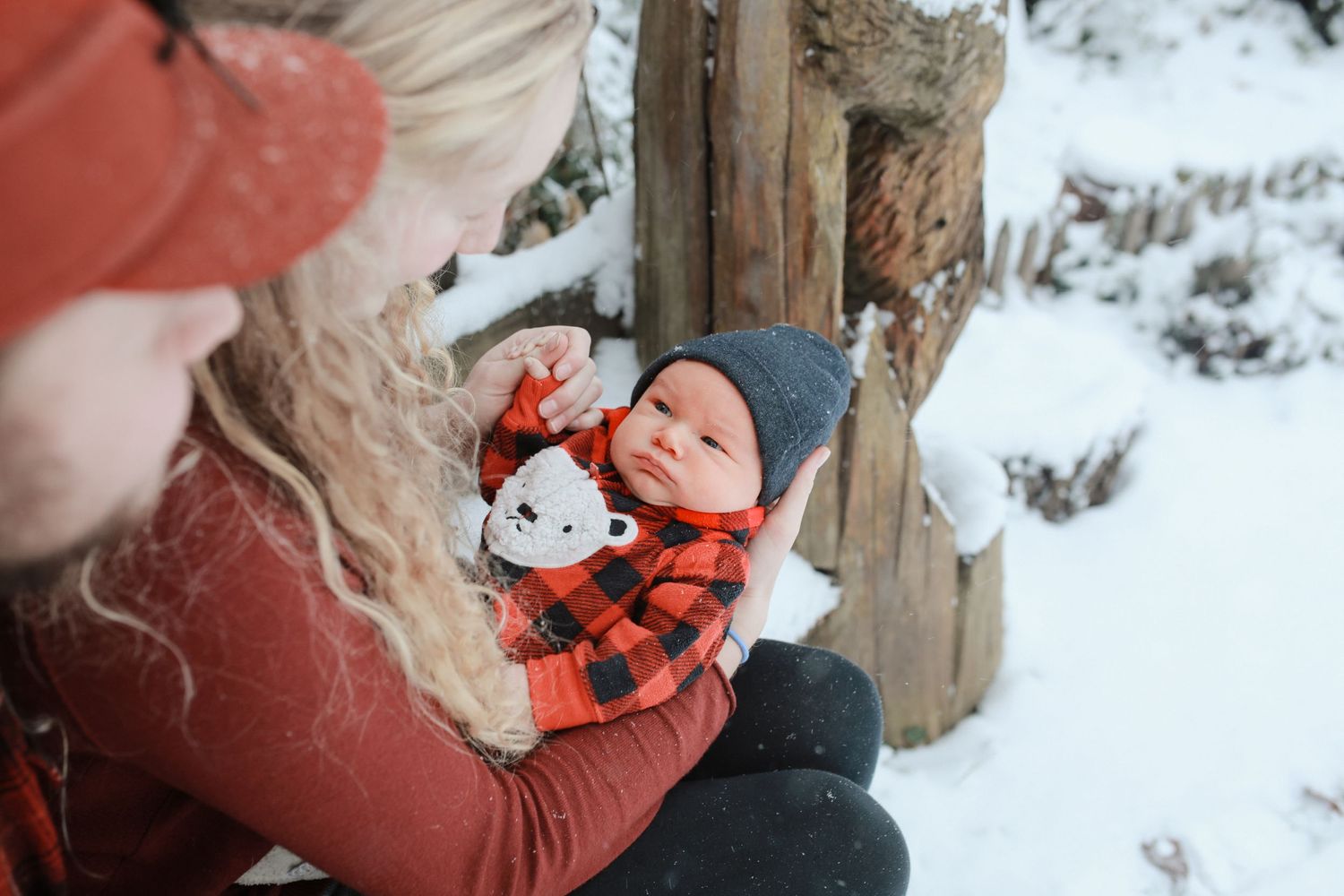 Mother tenderly holds her baby boy over her knees, with the baby in a cozy red fuzzy outfit and grey hat. They are surrounded with snow.