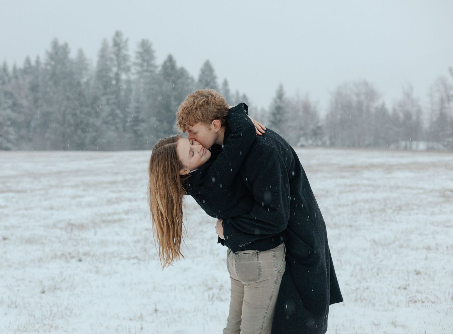 Romantic winter kiss in a snow-covered field, a couple embracing with joy and love amidst softly falling snowflakes and a serene forest back