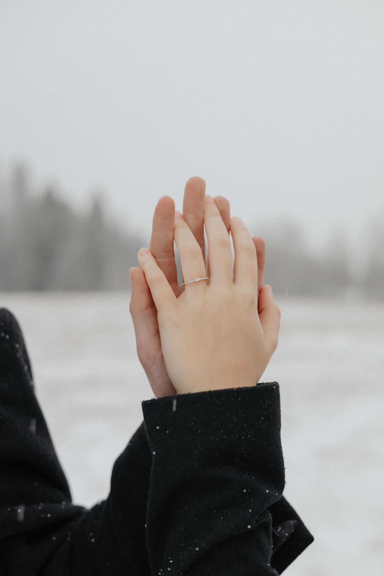 A close-up of a newly engaged couple’s hands clasped together in a snowy landscape. The front-facing hand has an engagement ring on it.