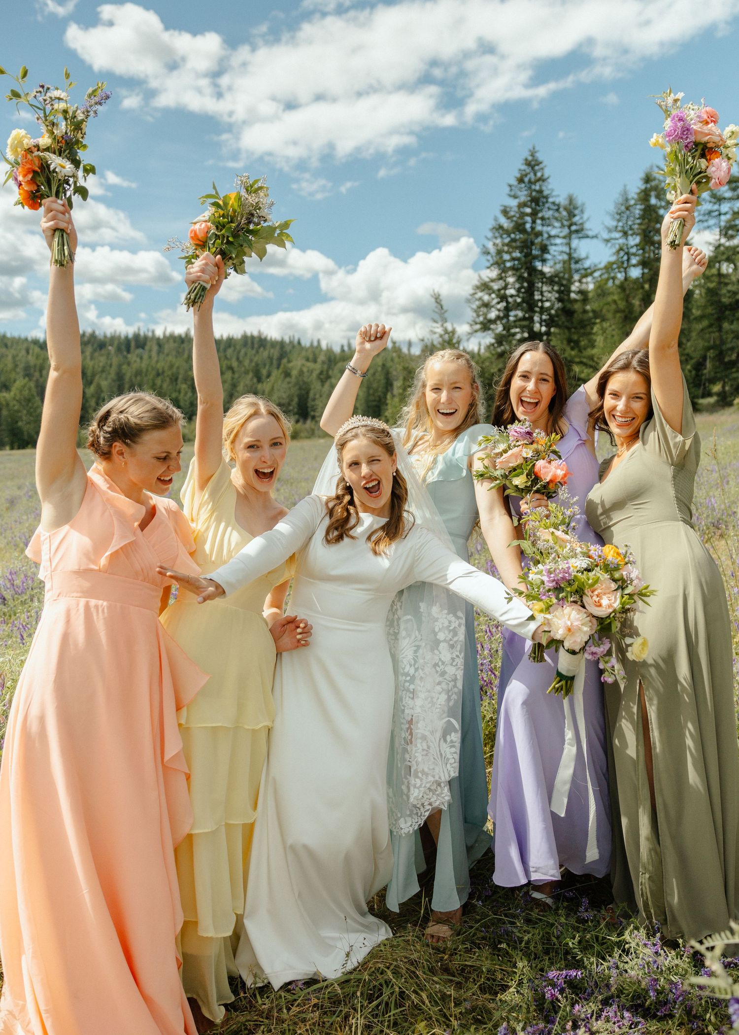 Colorful bridesmaids standing beside the bride in a field, holding up their bouquets in the air and cheering, with trees in the distance.