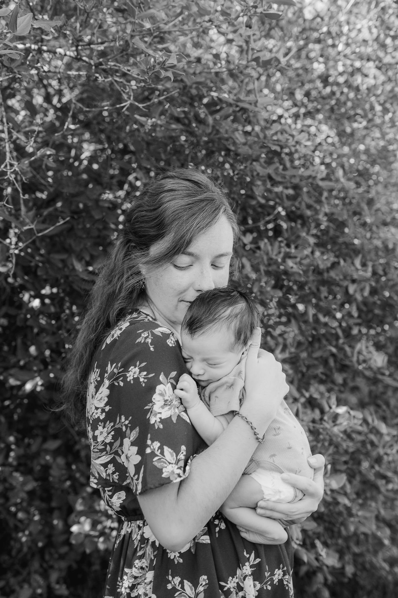 Black and White image of a mother holding her newborn baby with lush greenery behind her.
