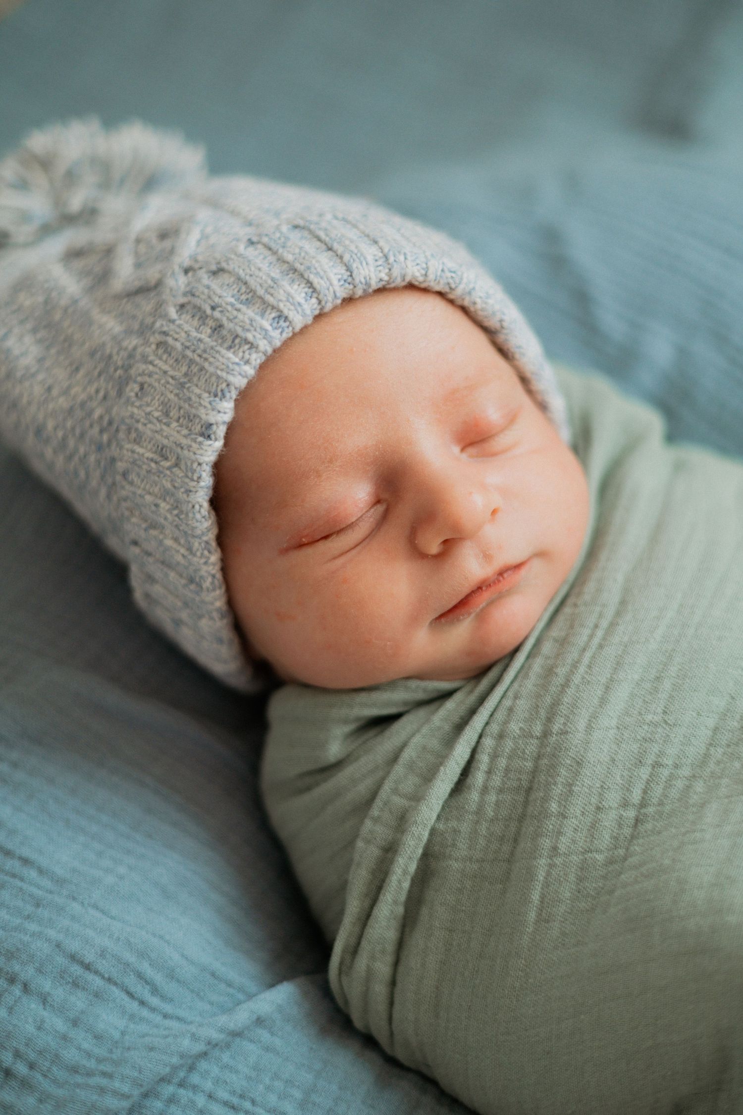 A newborn baby swaddled in a soft green blanket, wearing a gray knit hat, resting on a light blue blanket.