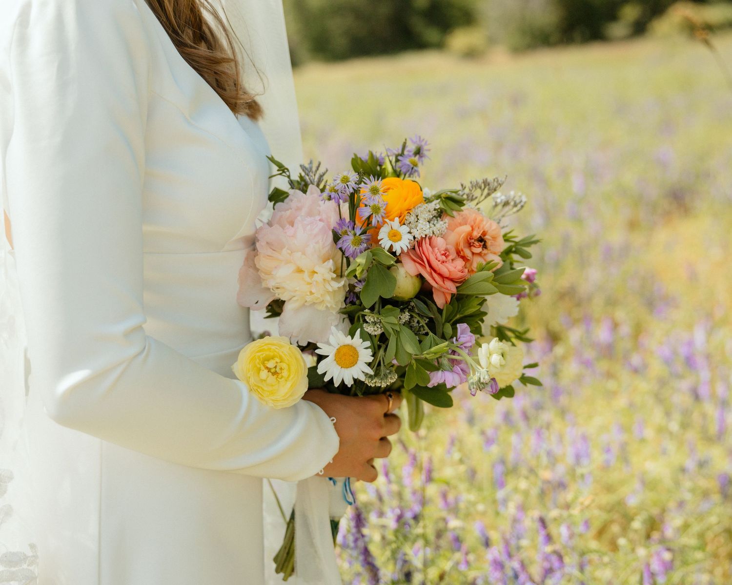 Bride gracefully holding a vibrant bouquet of colorful flowers, set against a field of purple blossoms in soft, natural light.