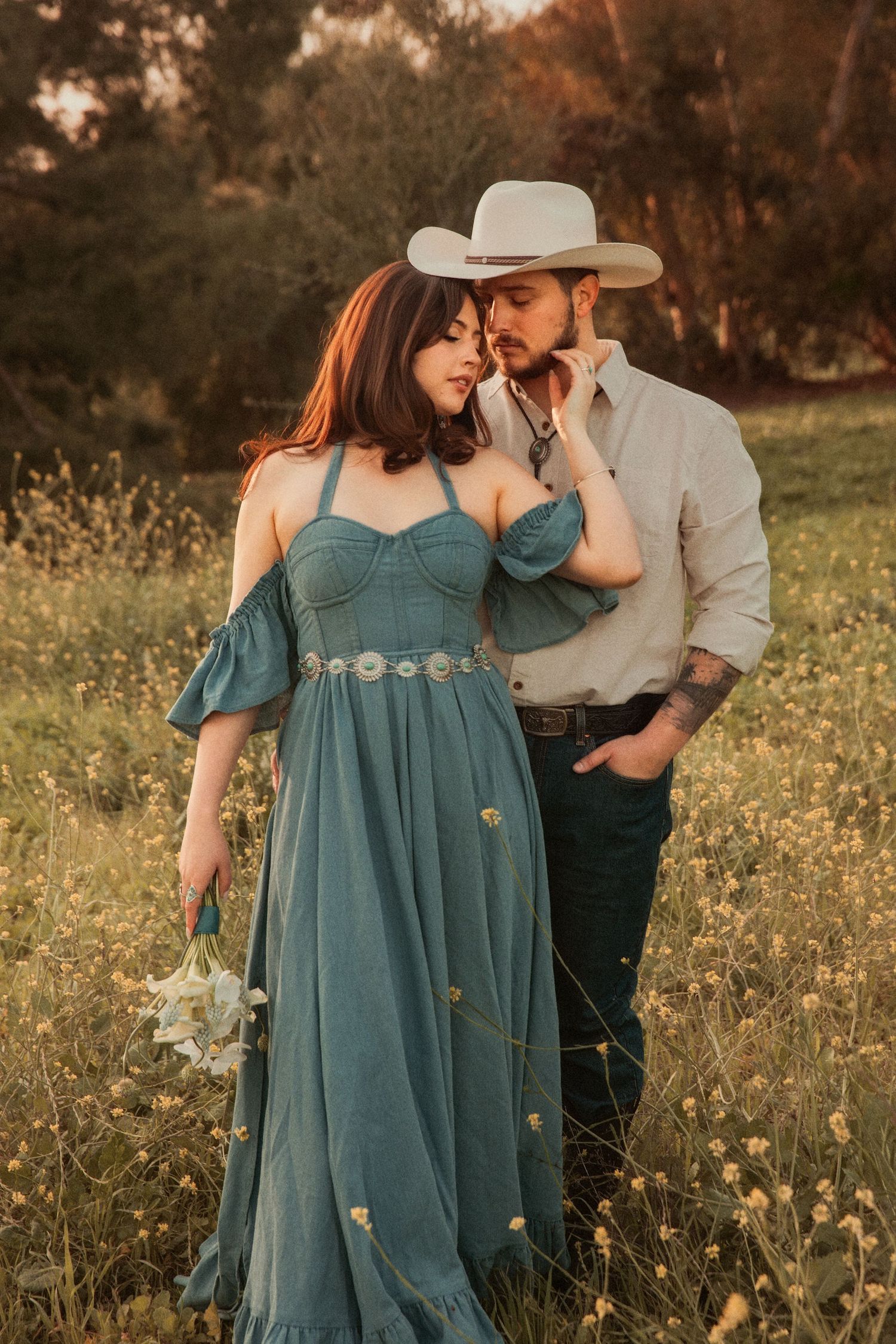 A romantic sunset portrait in a wildflower meadow with a cowboy hat and flowing teal dress capturing an intimate moment.