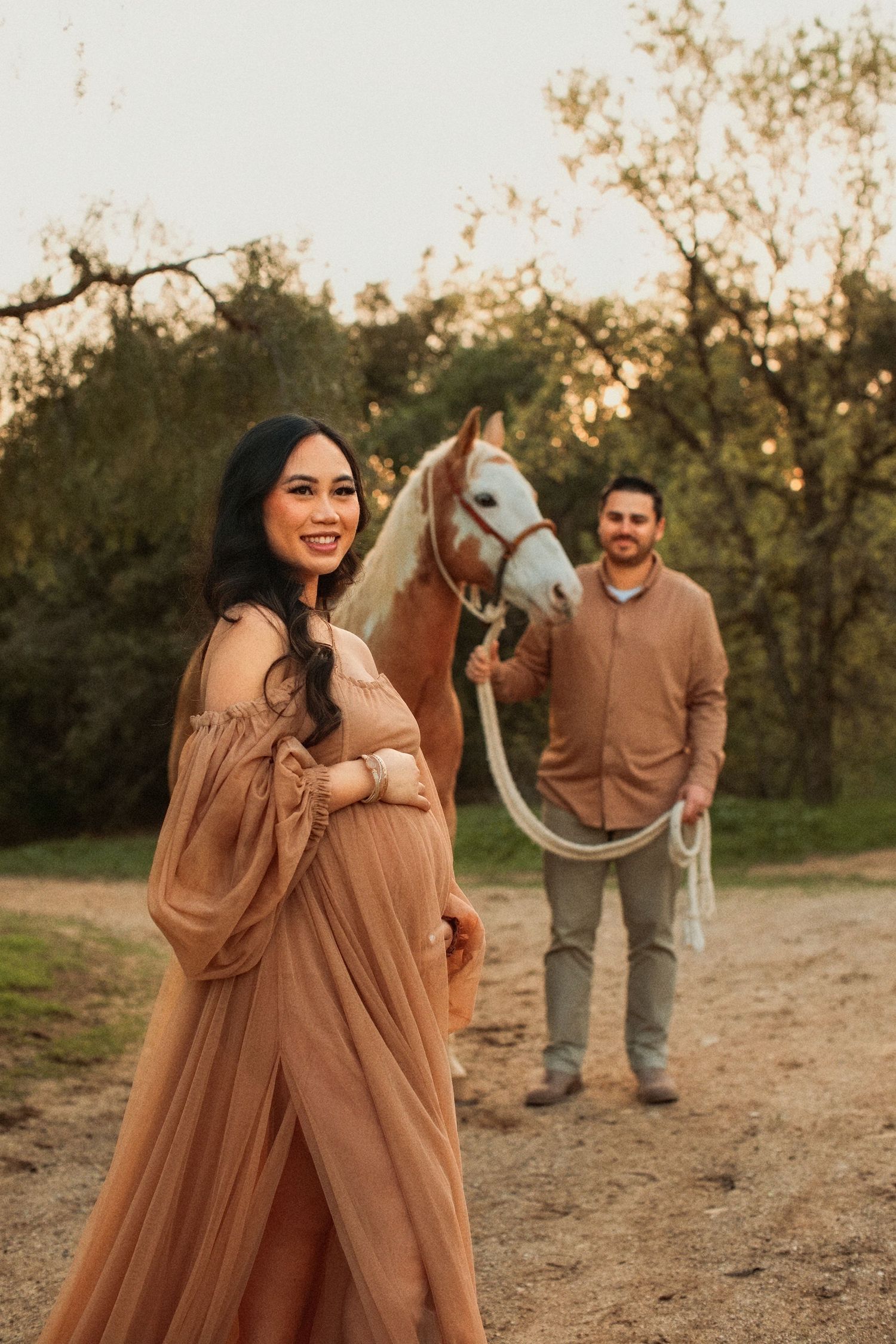 Outdoor maternity photo at sunset with a horse standing between a woman in a flowy peach dress and a man in earth tones.