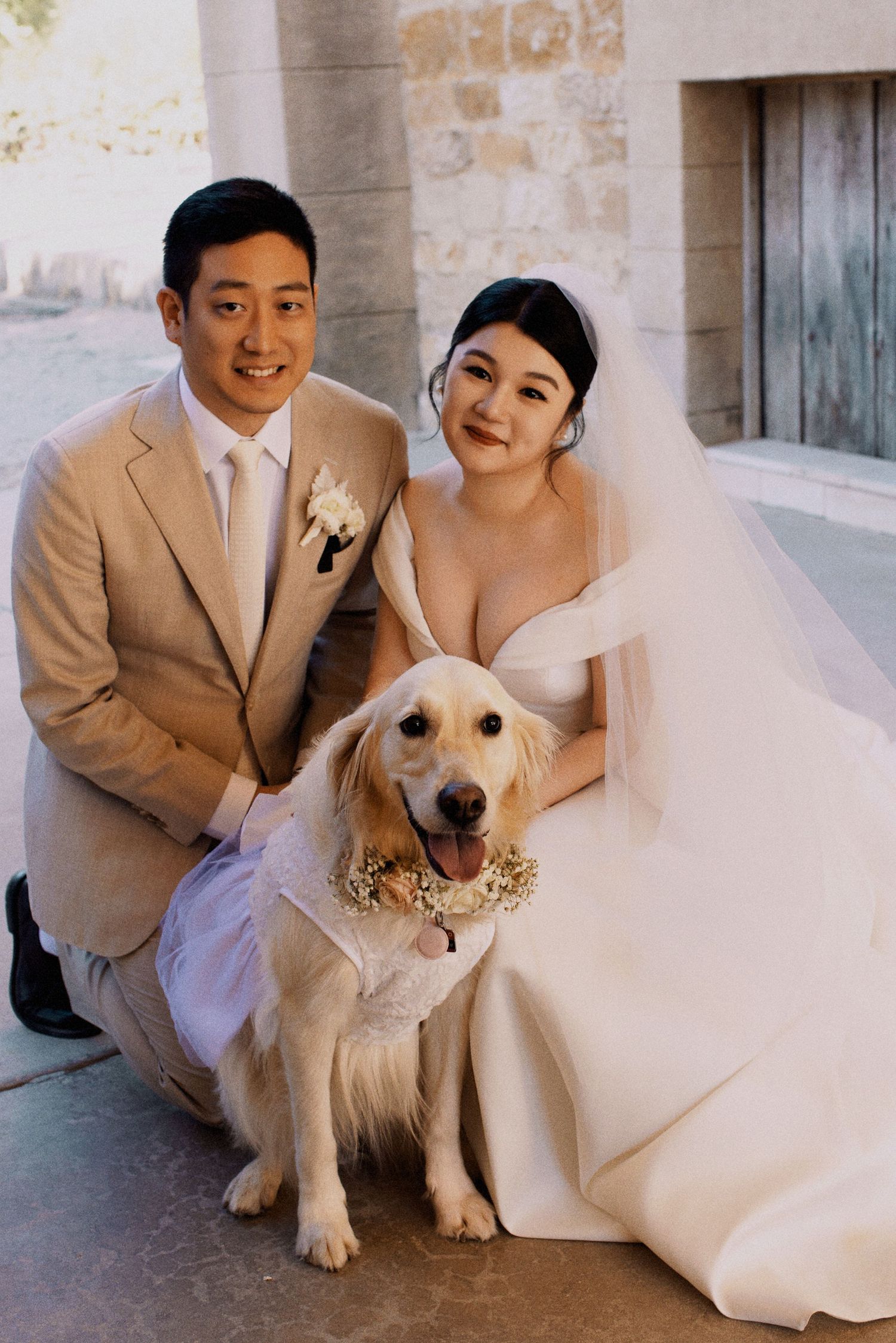 A wedding couple poses with their golden retriever who is wearing a floral collar at their ceremony.