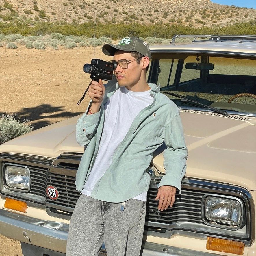 Someone takes photos with a camera while leaning against a vintage tan-colored car in a desert setting.