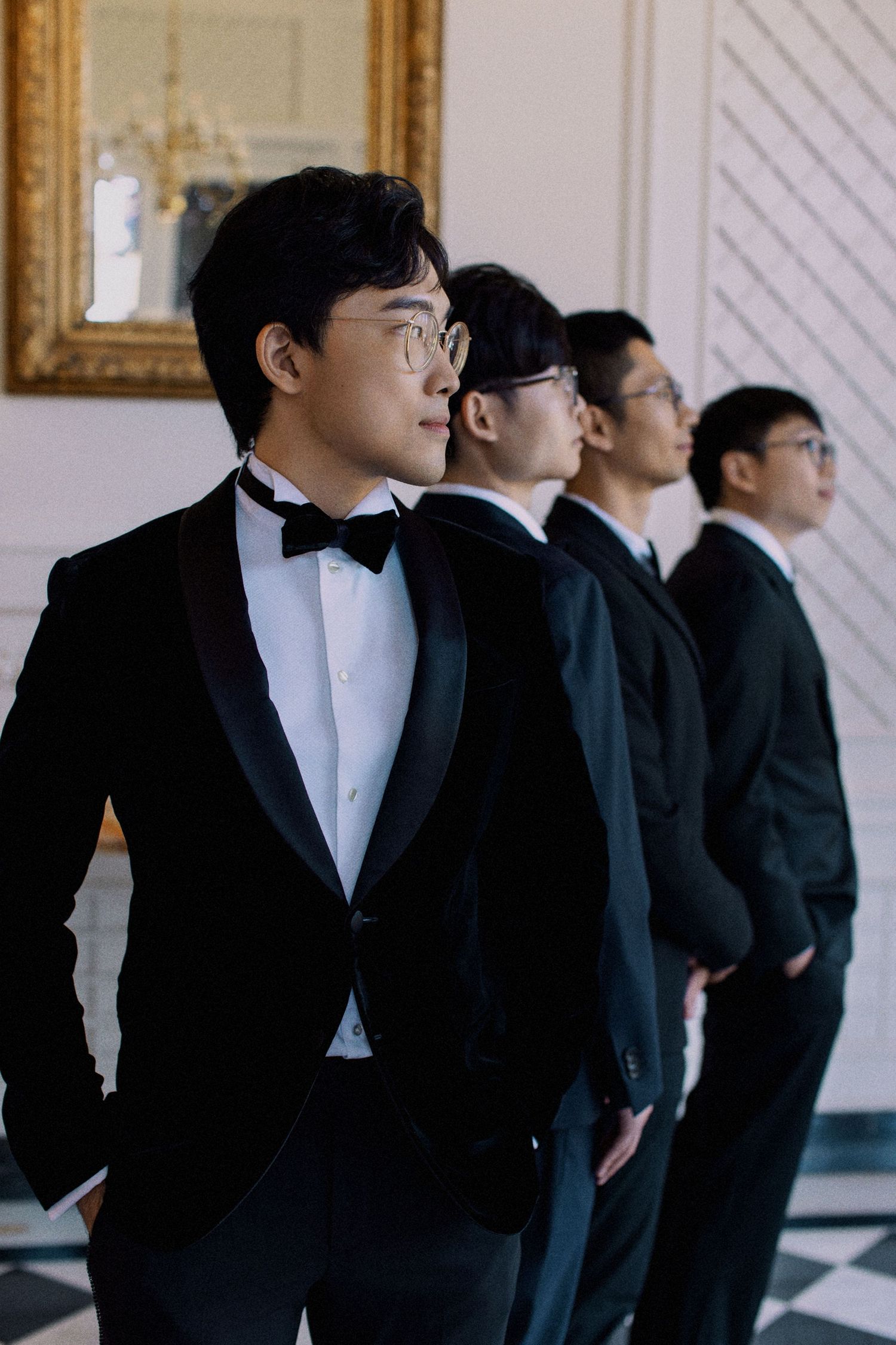 Groomsmen in formal black tuxedos and bow ties lined up against a white wall.