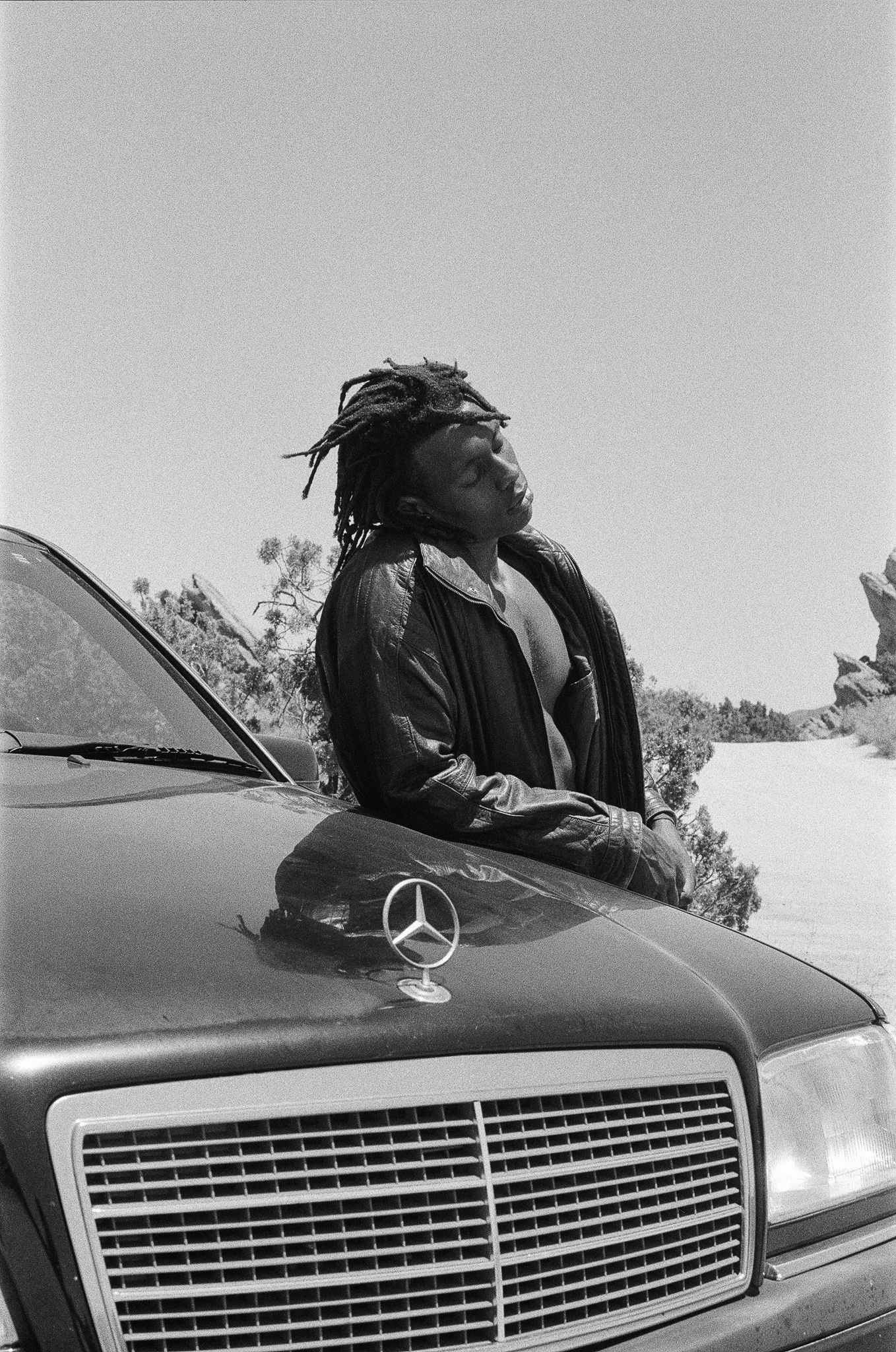 Black and white photo of person sitting on Mercedes-Benz hood wearing leather jacket.