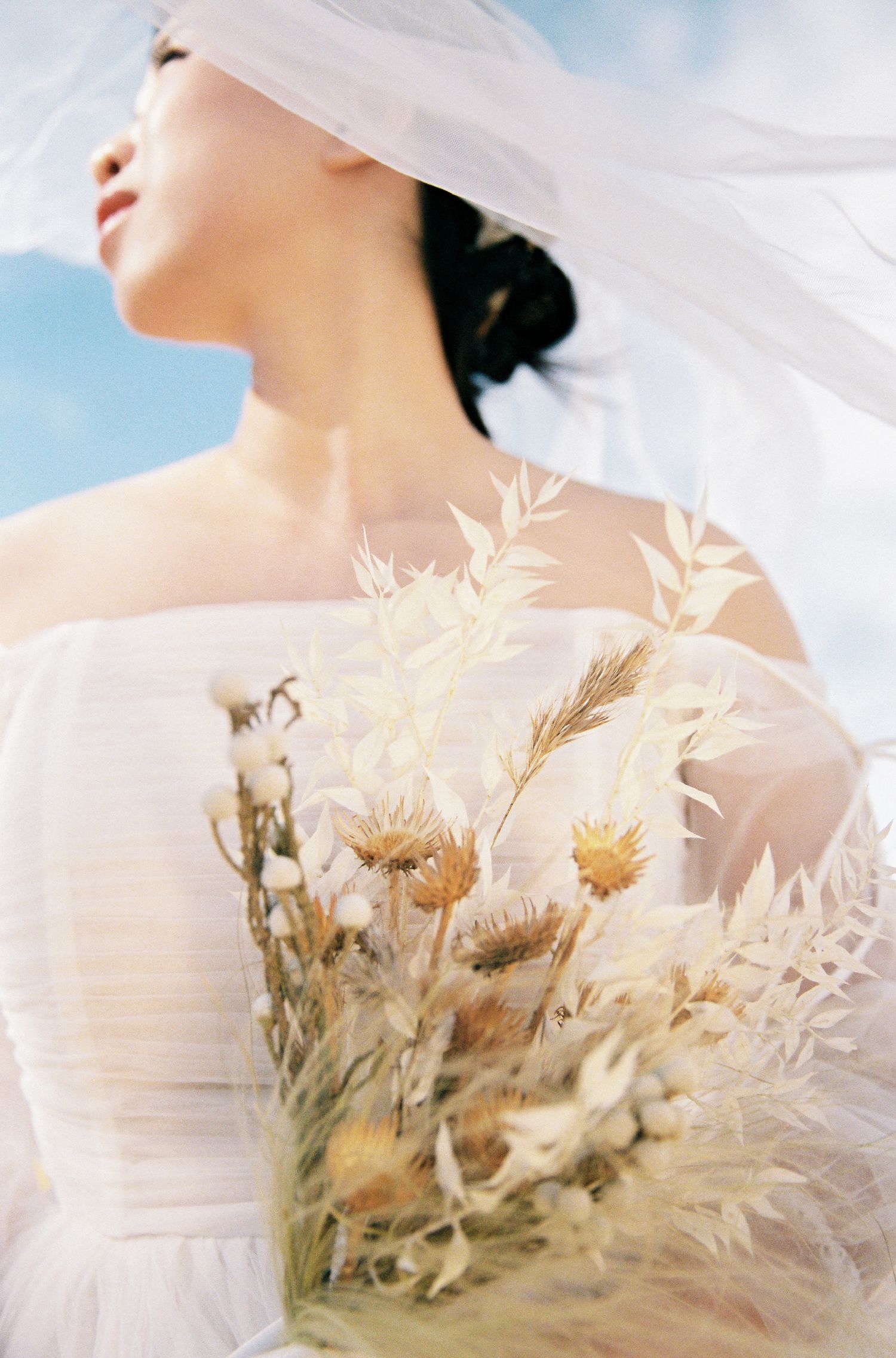 Ethereal bridal portrait with dried flowers against blue sky.