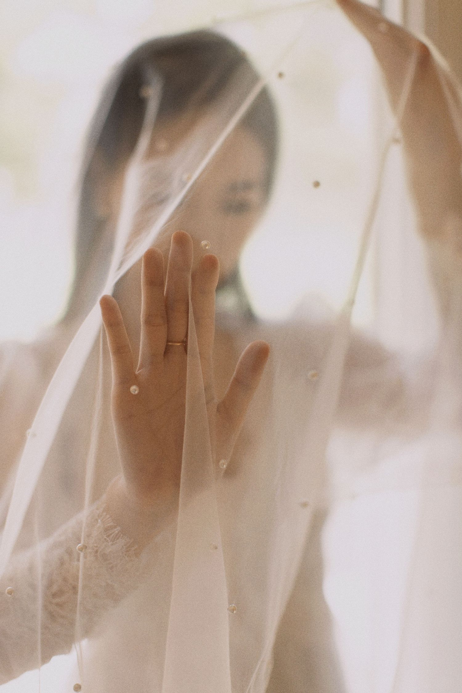 An artistic bridal portrait showing a hand with engagement ring through sheer wedding veil.
