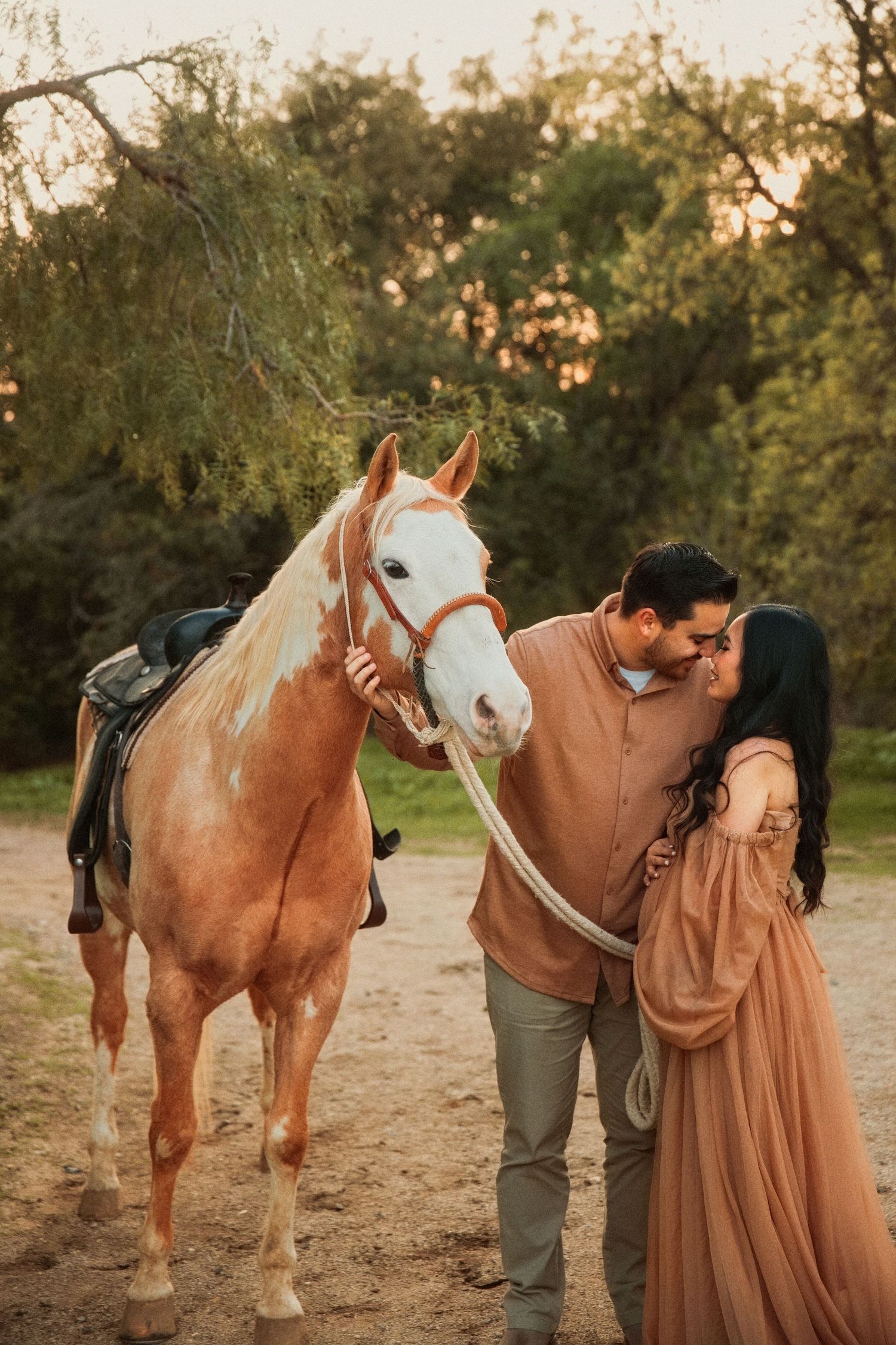 A beautiful palomino horse stands on a dirt path at sunset while a couple shares an intimate moment beside it.