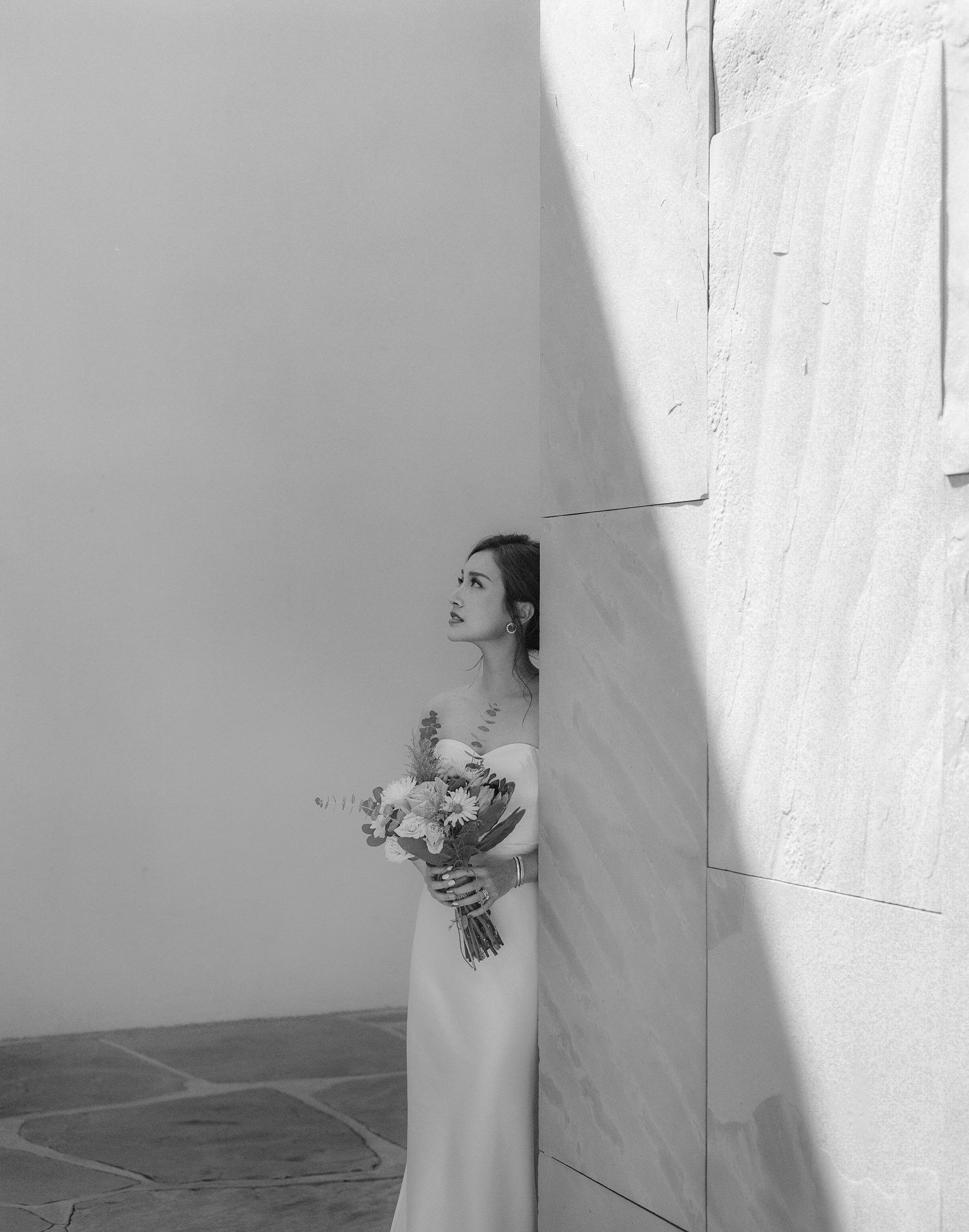 Black and white photo of bride holding bouquet against white architectural wall.