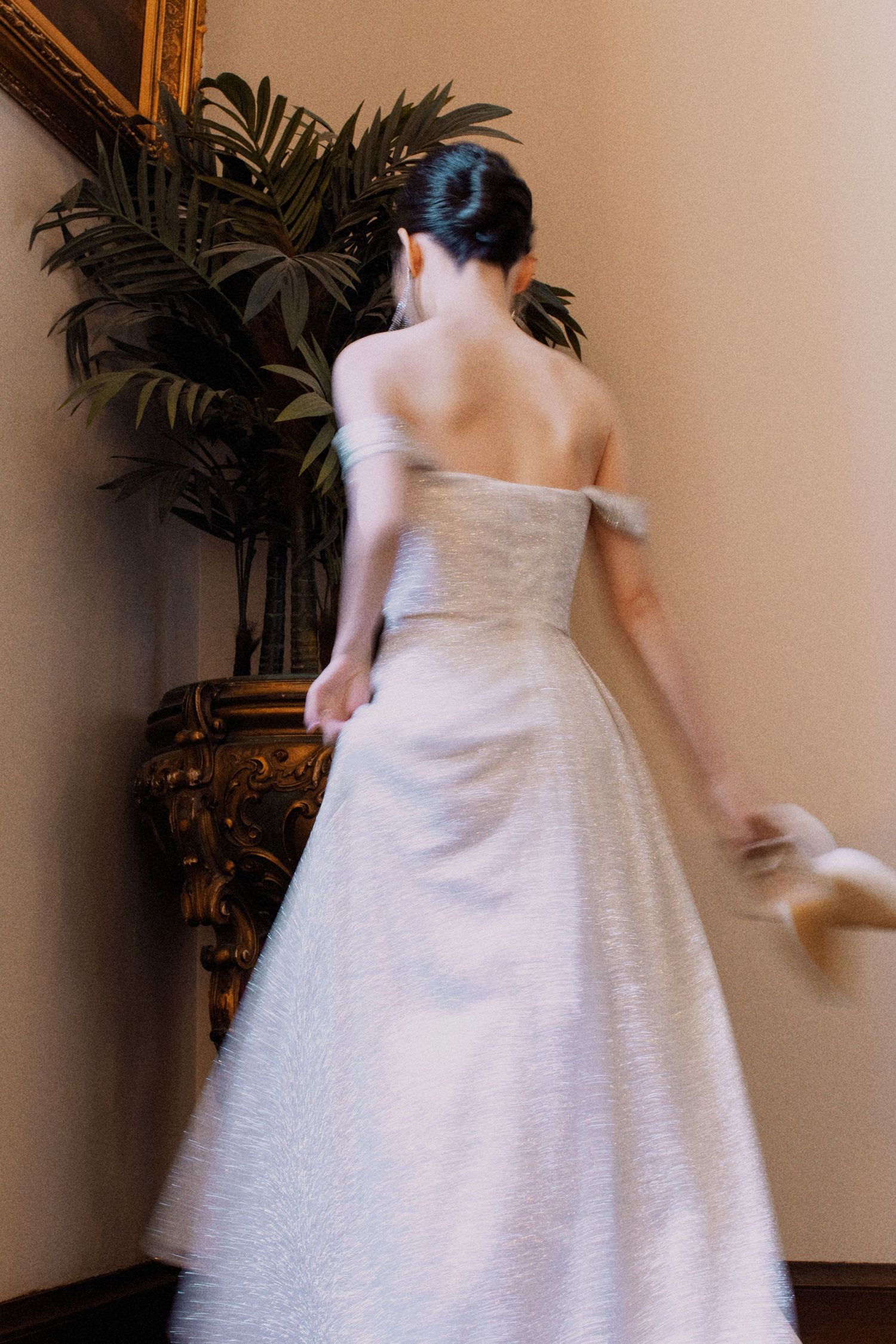 Back view of a bride in an elegant off-shoulder white wedding gown standing near decorative plants.