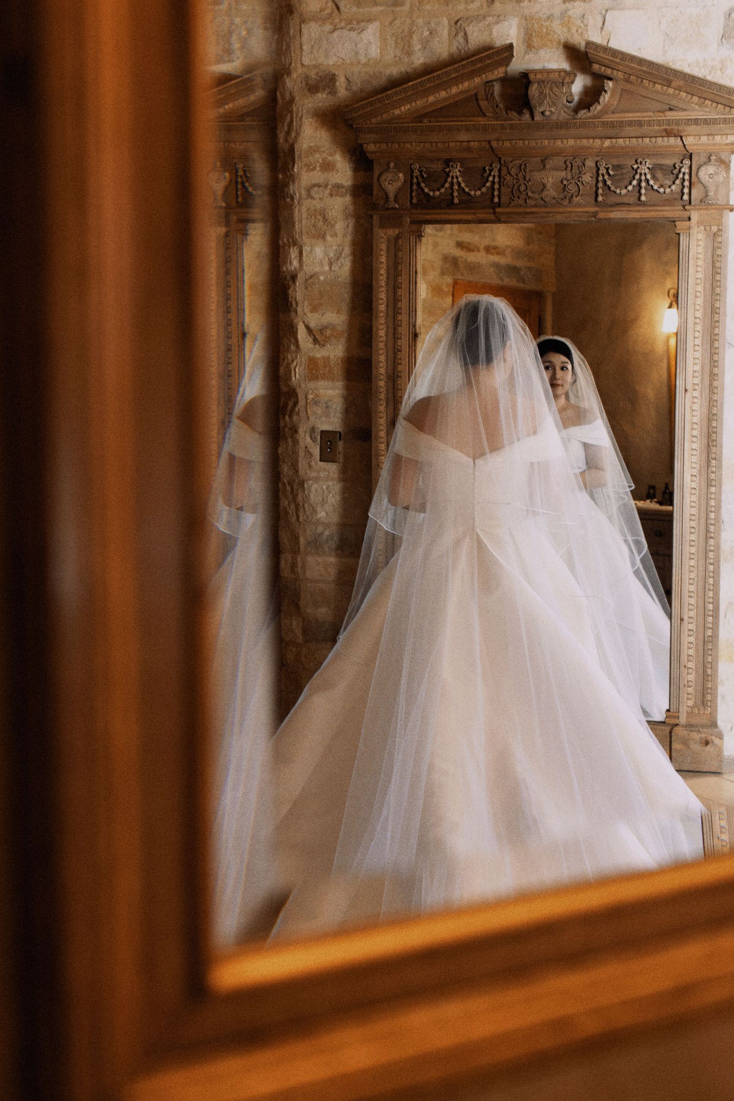 A bride's reflection in an ornate vintage mirror shows her flowing white veil and wedding gown.