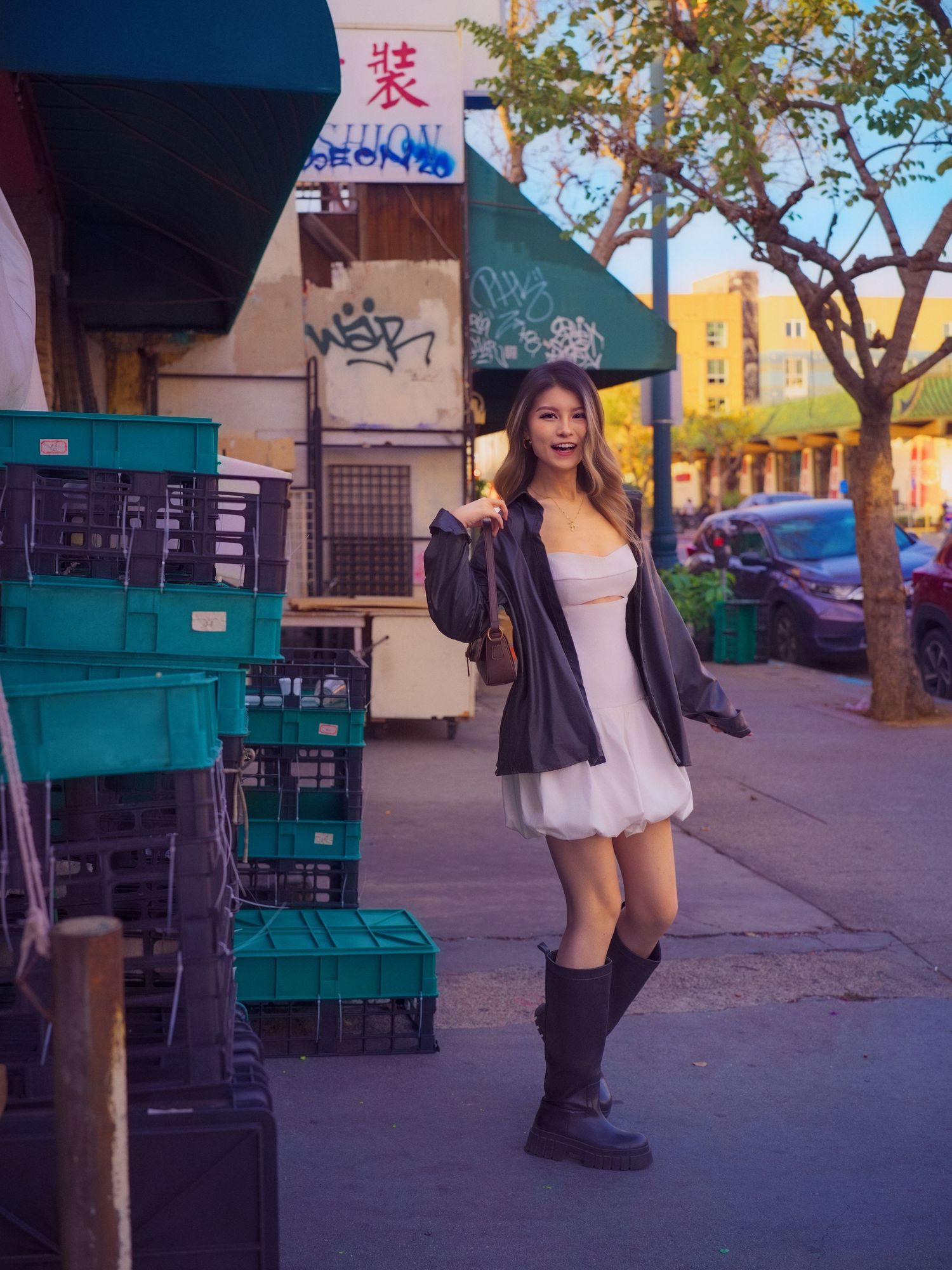 Model in white dress and boots poses on urban sidewalk near green containers.