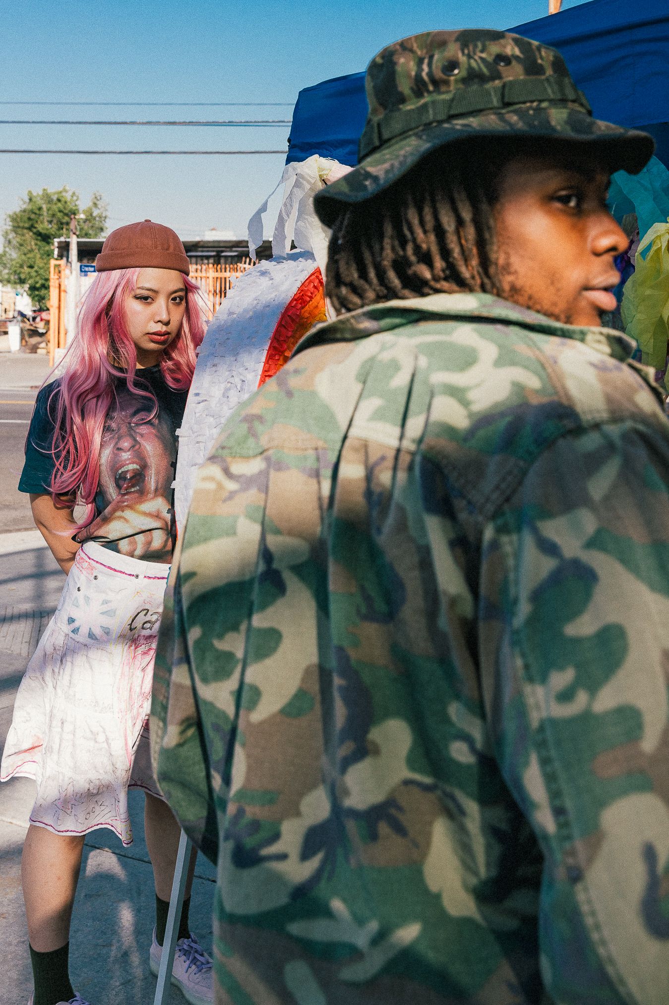 Urban fashion shoot with pink-haired model and person in camo gear.
