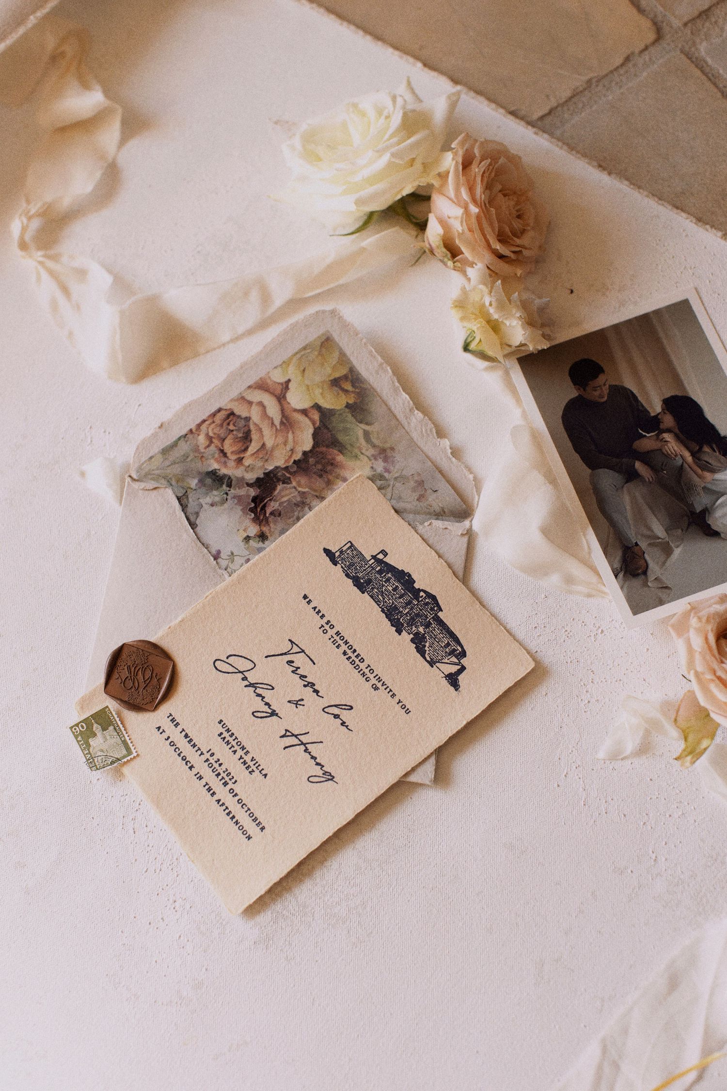 Vintage wedding invitation with wax seal and dried roses on white surface.