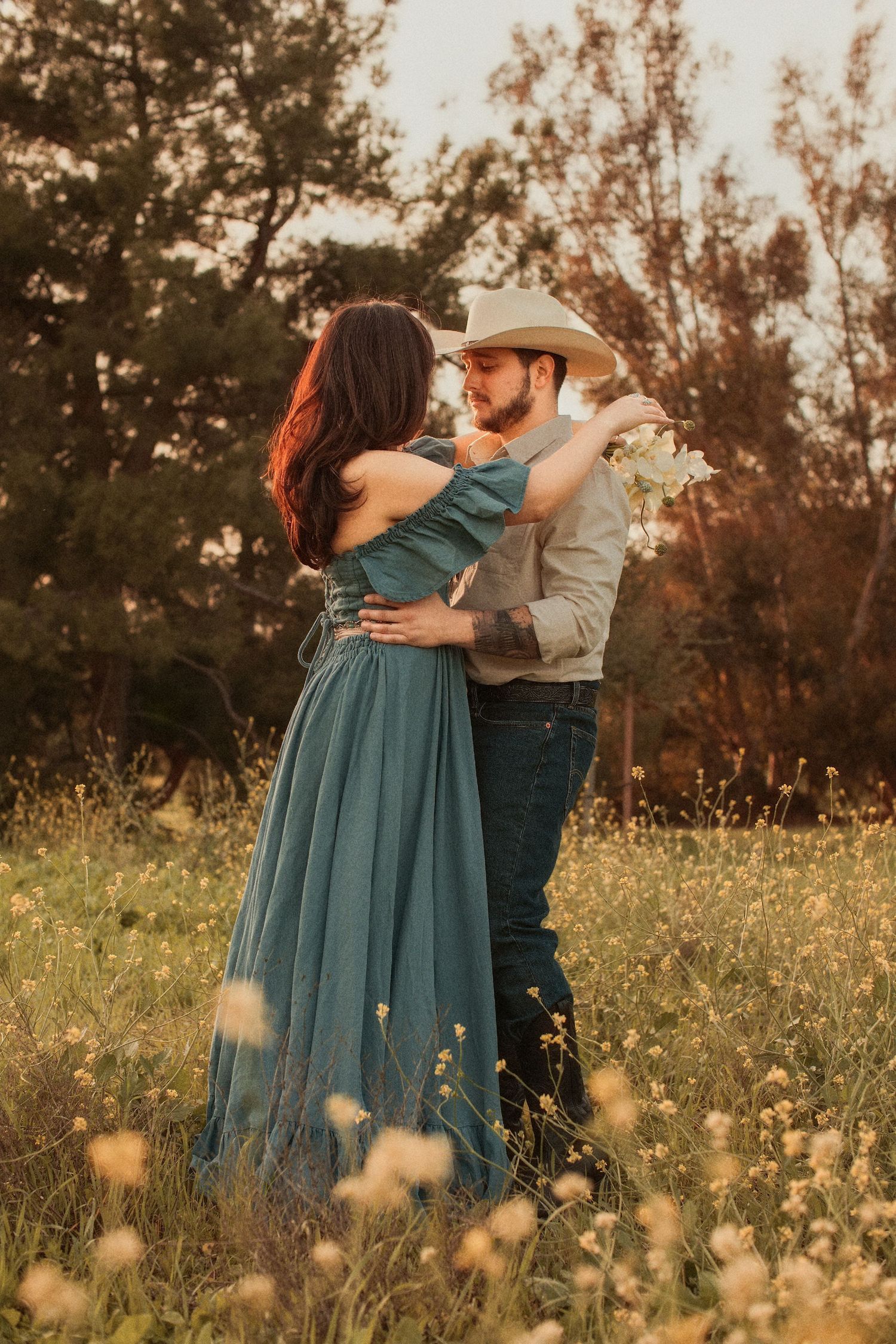 Romantic couple embraces in a wildflower meadow at sunset, with woman in teal dress and man wearing cowboy hat.