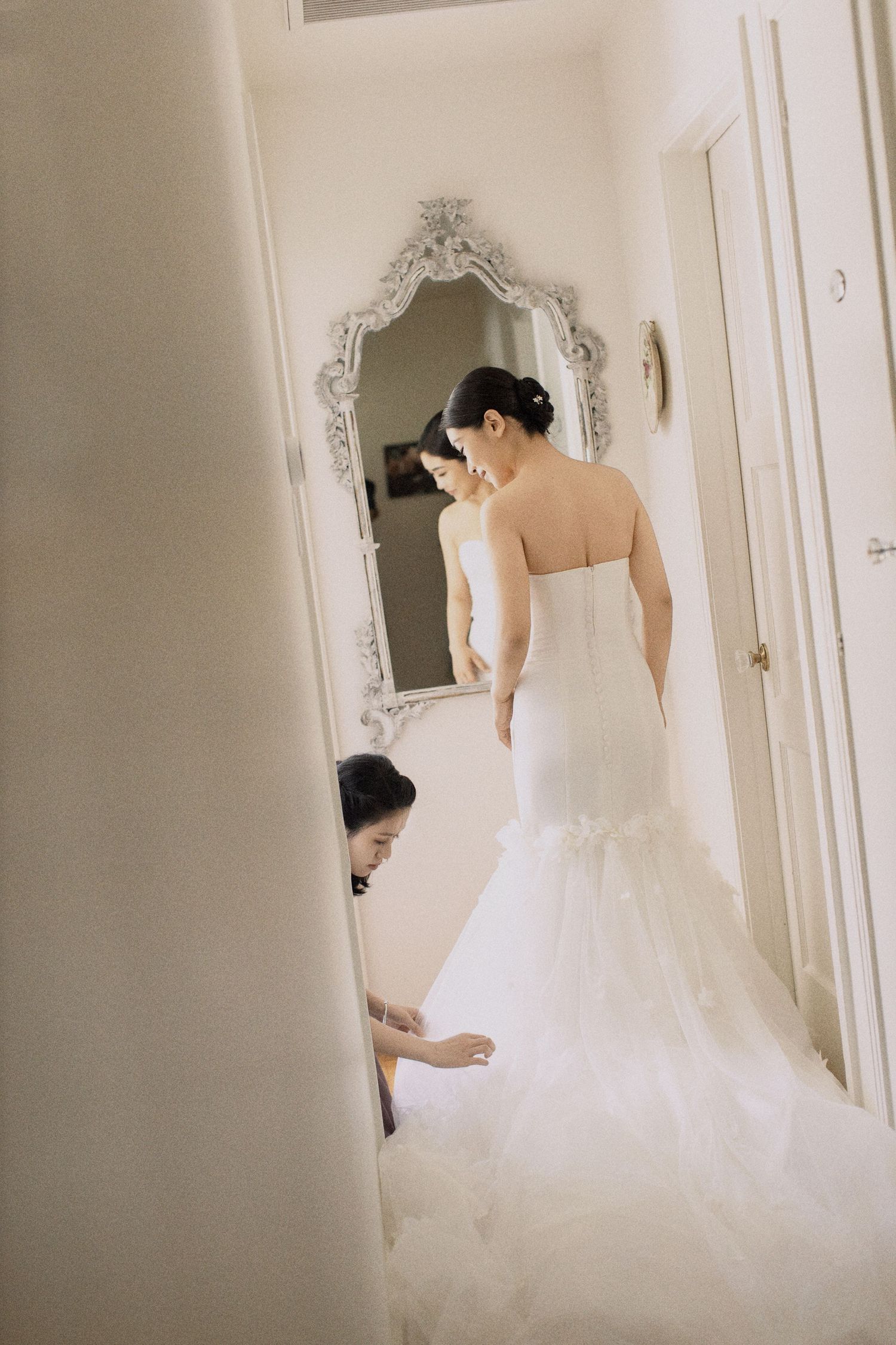 Wedding dress being adjusted in an elegant room with ornate mirror.