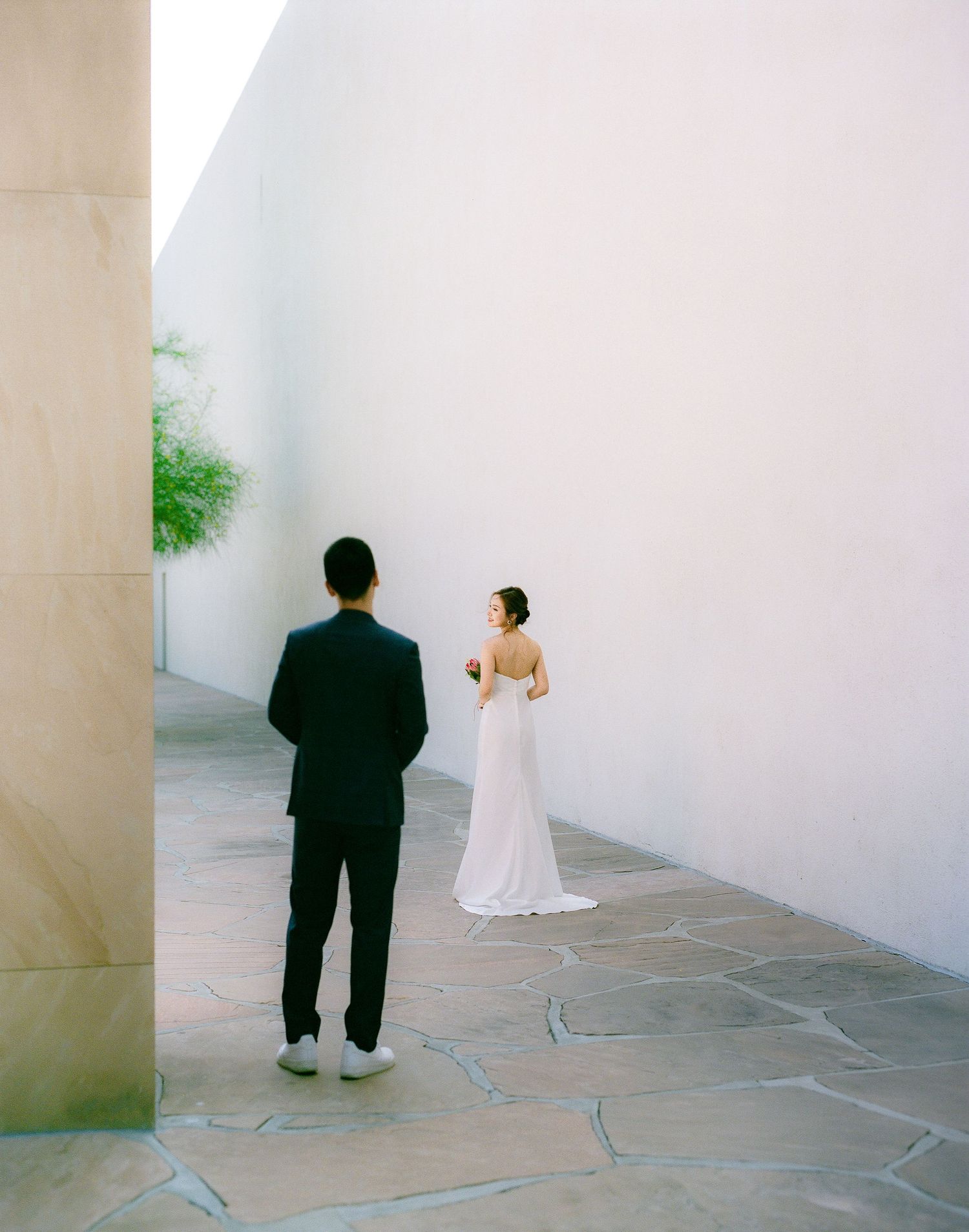 A couple in wedding attire stand apart on stone pavement with white walls.